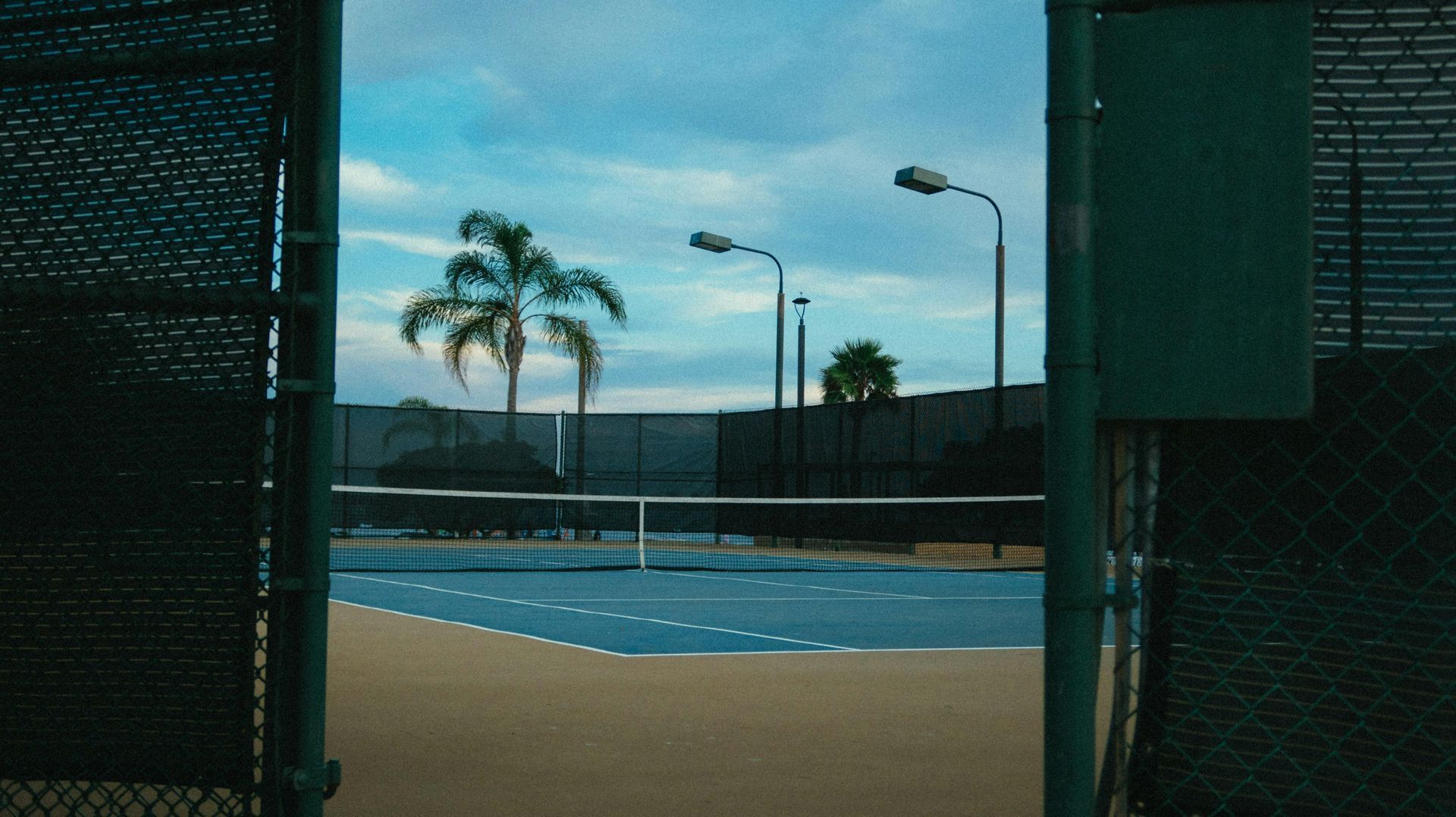 Open gate view of a blue tennis court with palm trees and lampposts under a cloudy sky.