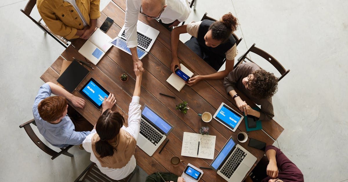 People at a wooden table shaking hands, laptops and tablets present, overhead view.