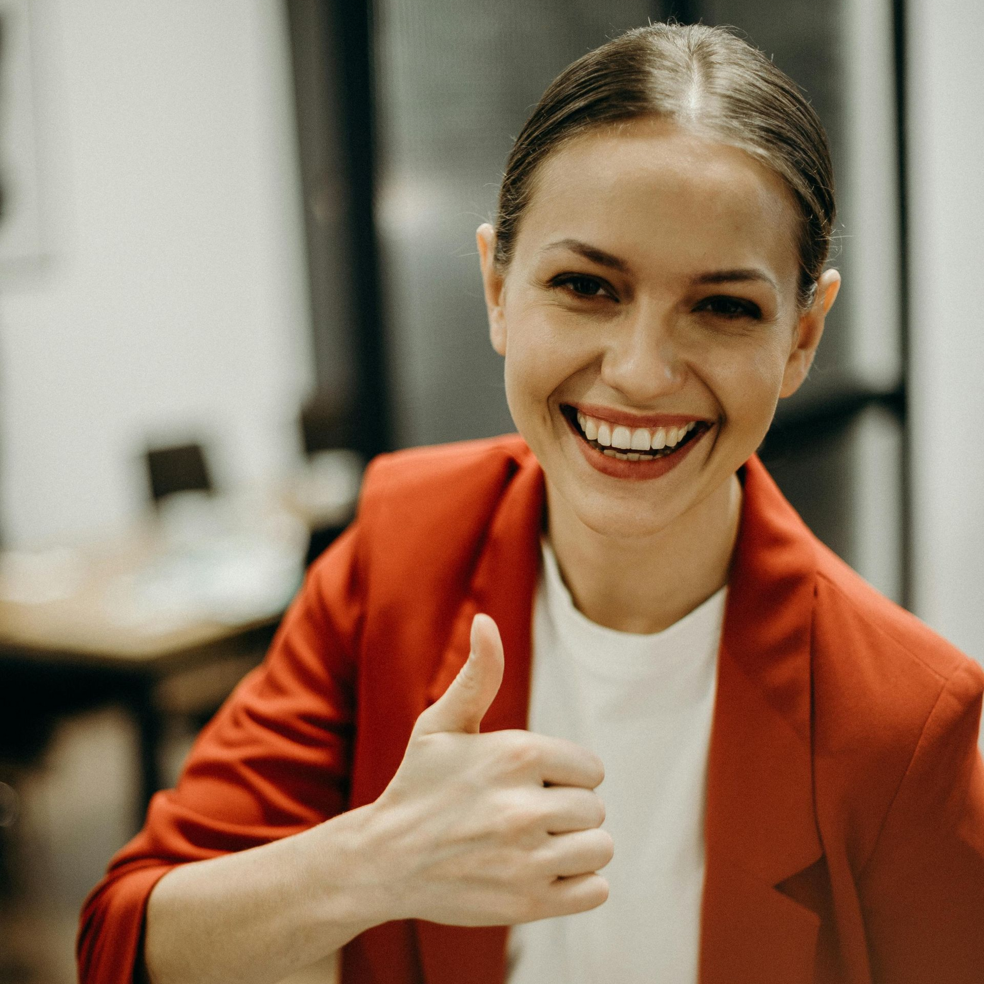 Woman in red blazer smiles, giving a thumbs up. Office setting.