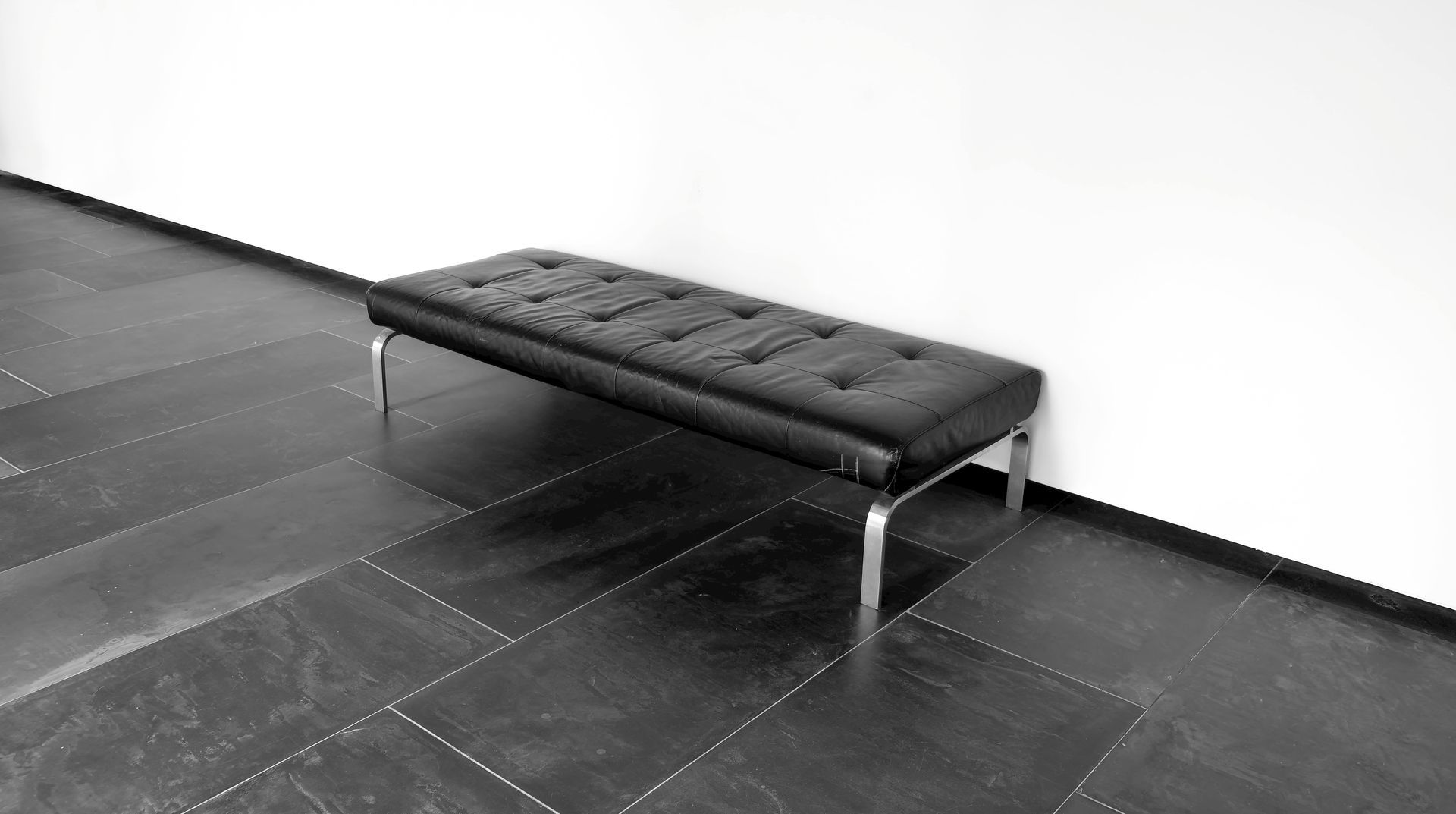 Black and white photo: A tufted bench with metal legs sits against a white wall on a tiled floor.