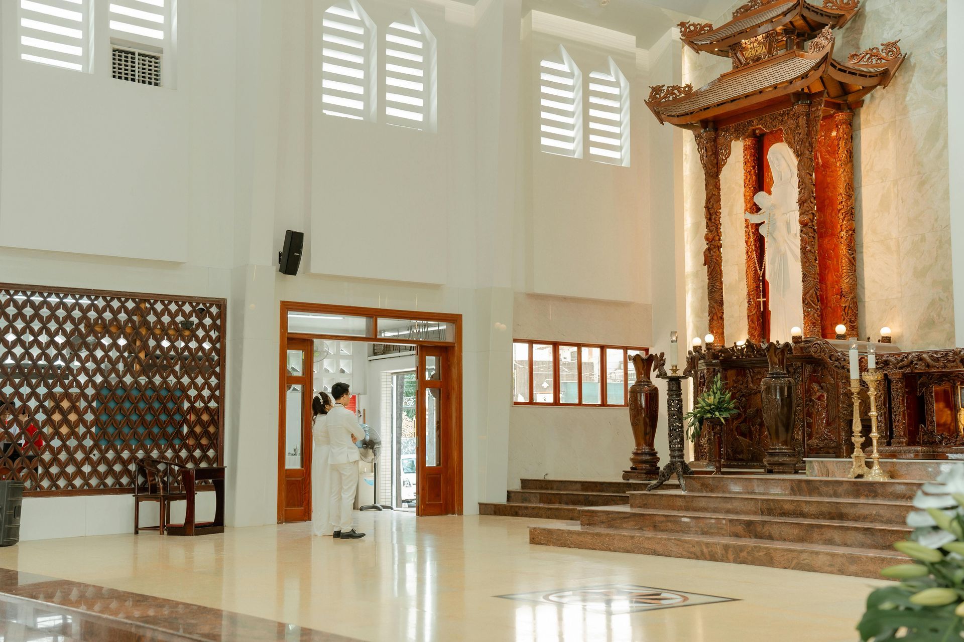 Priest in white robes stands at doorway of a bright church with ornate altar.