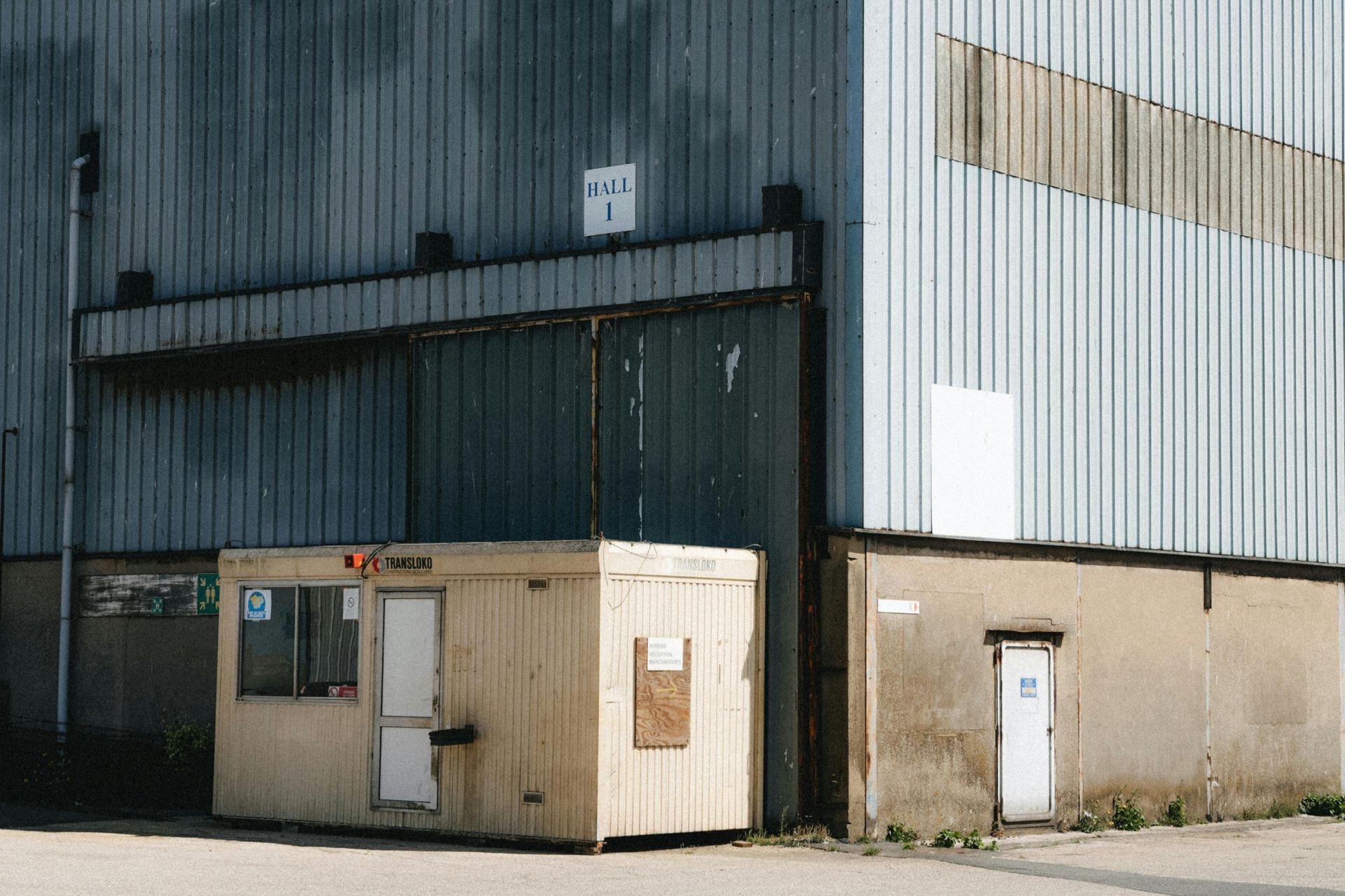 A small wooden office next to a large industrial building with a small door and a sign.