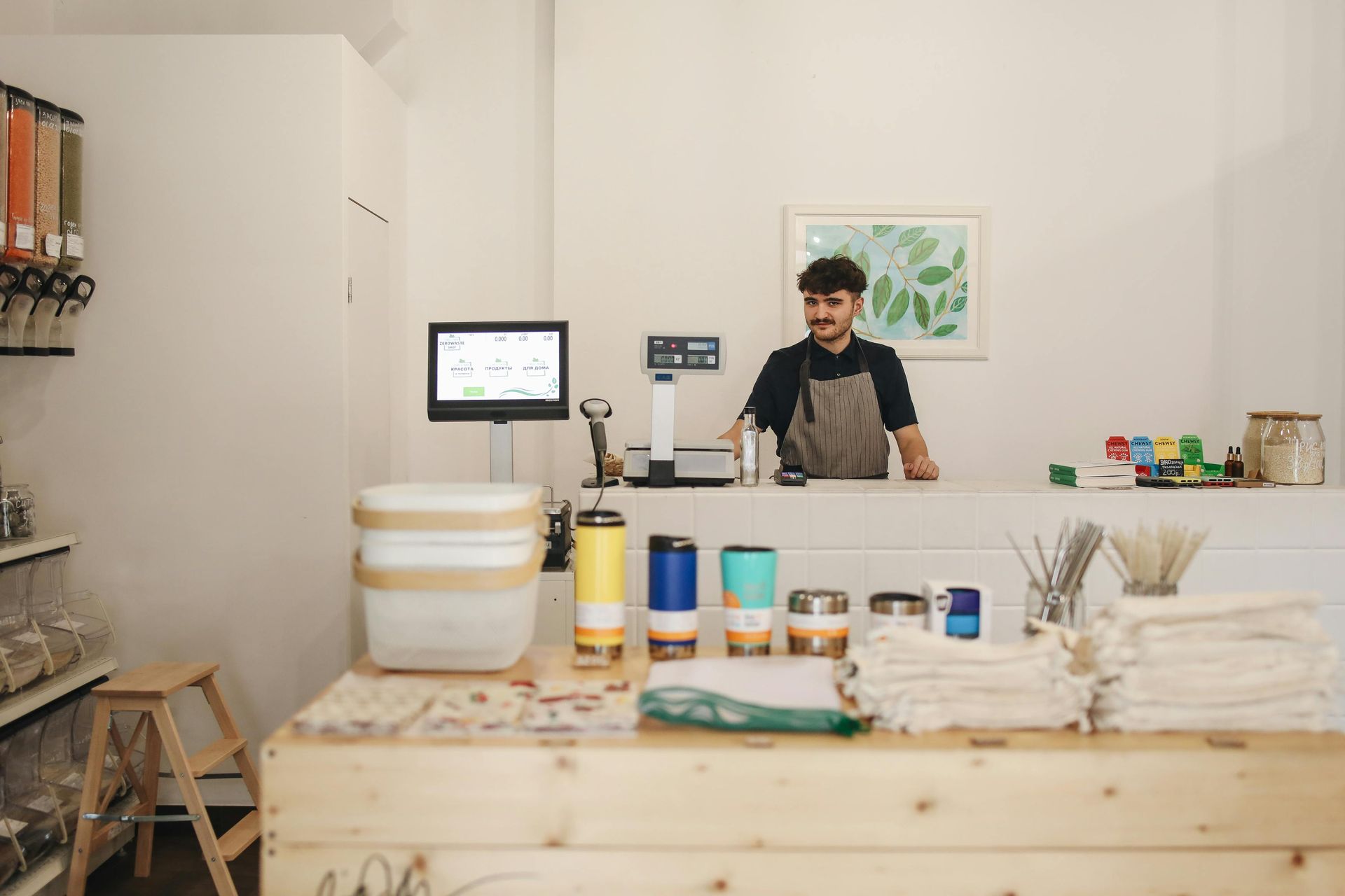 Man behind counter at zero-waste store. Smiling, wearing apron. Counter has containers, scales, reusable items.