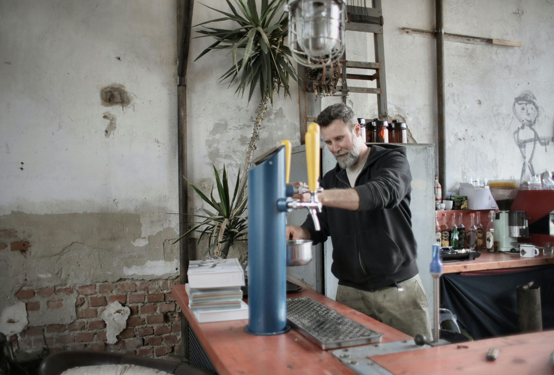 Man pours beer from tap at a rustic bar. He's smiling. The setting has exposed brick, a plant, and a ladder.