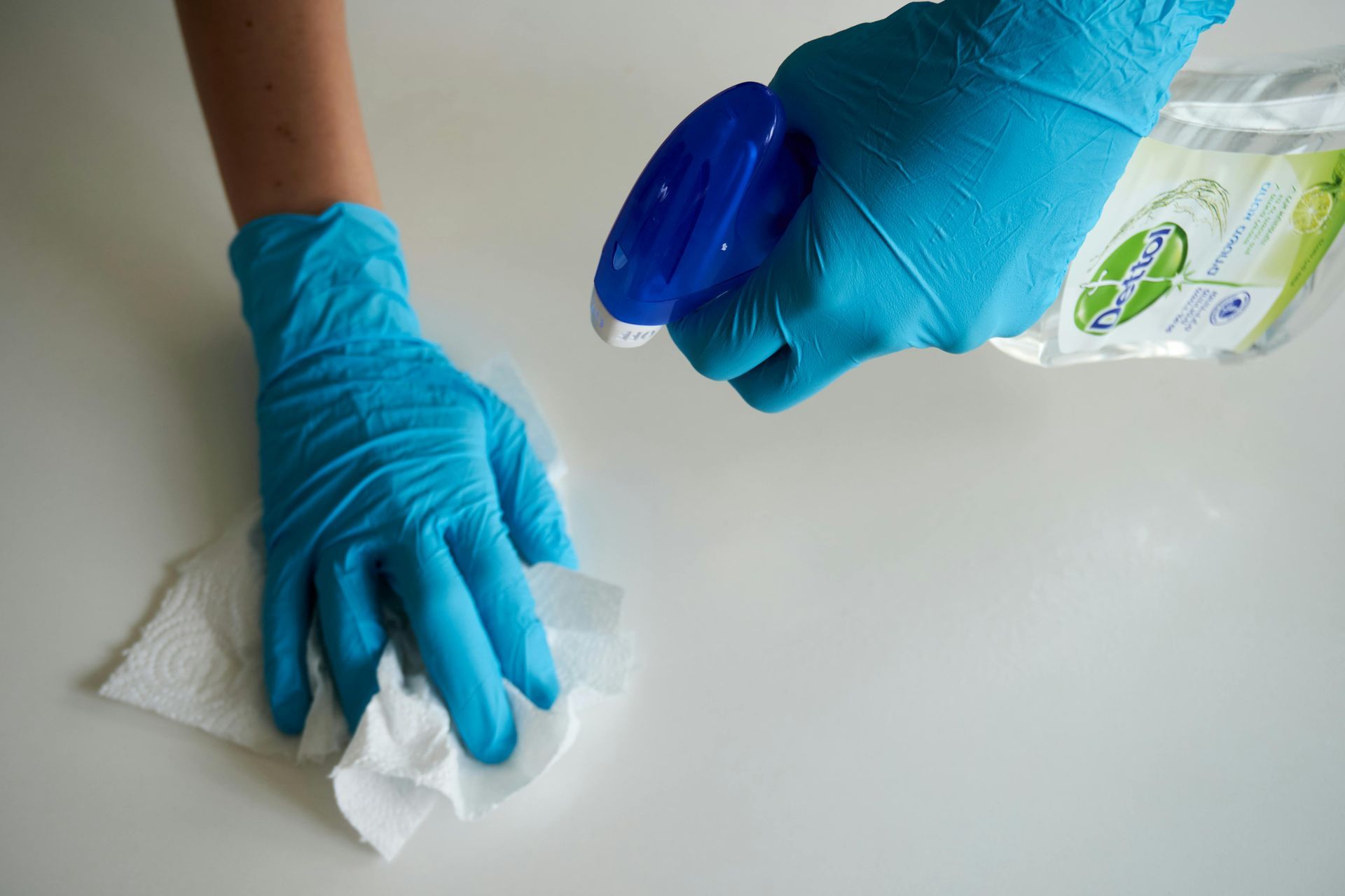 Hands in blue gloves spraying cleaner on a white surface and wiping with a paper towel.