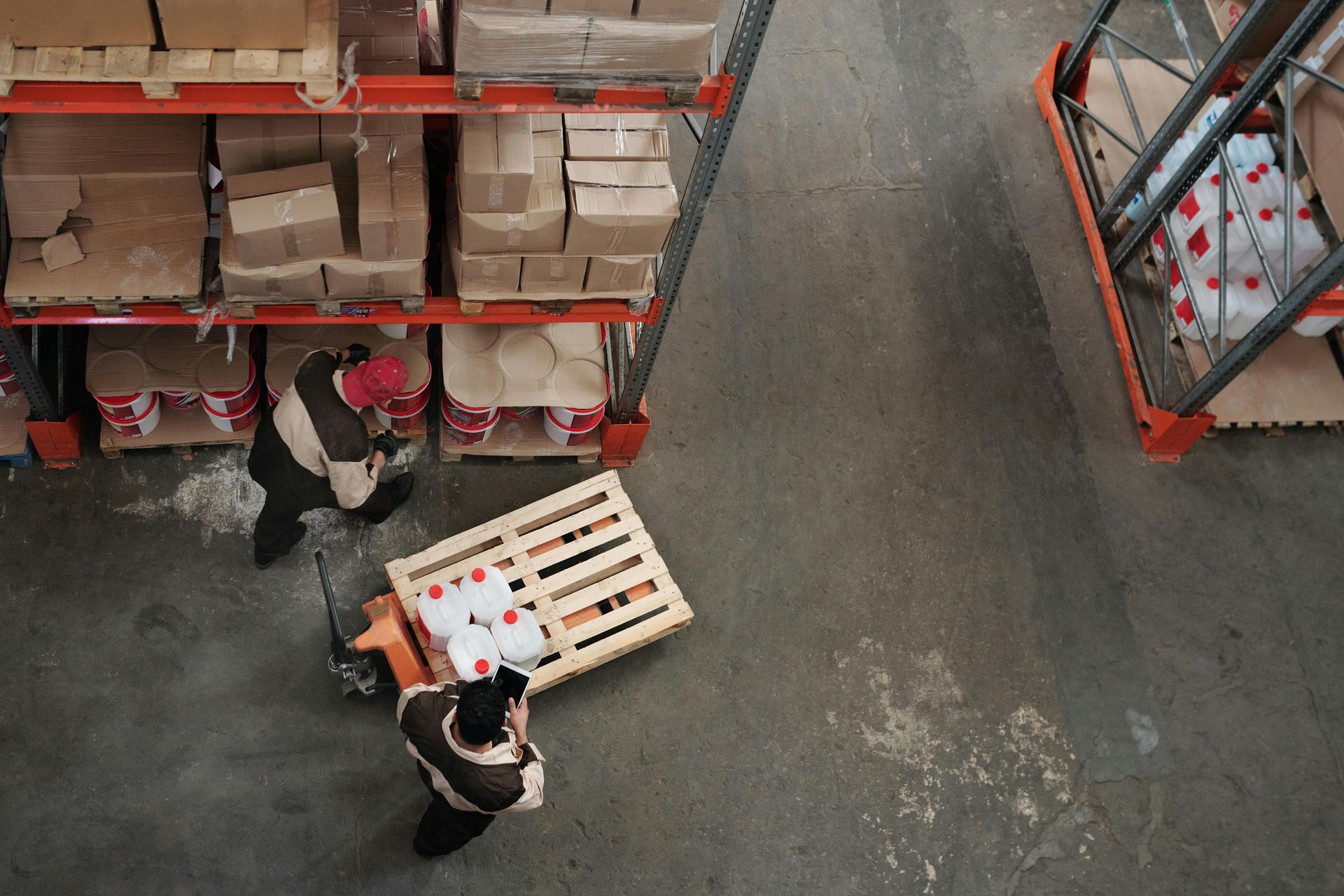 Overhead view of two people in warehouse, one using pallet jack to move wooden pallet of goods.