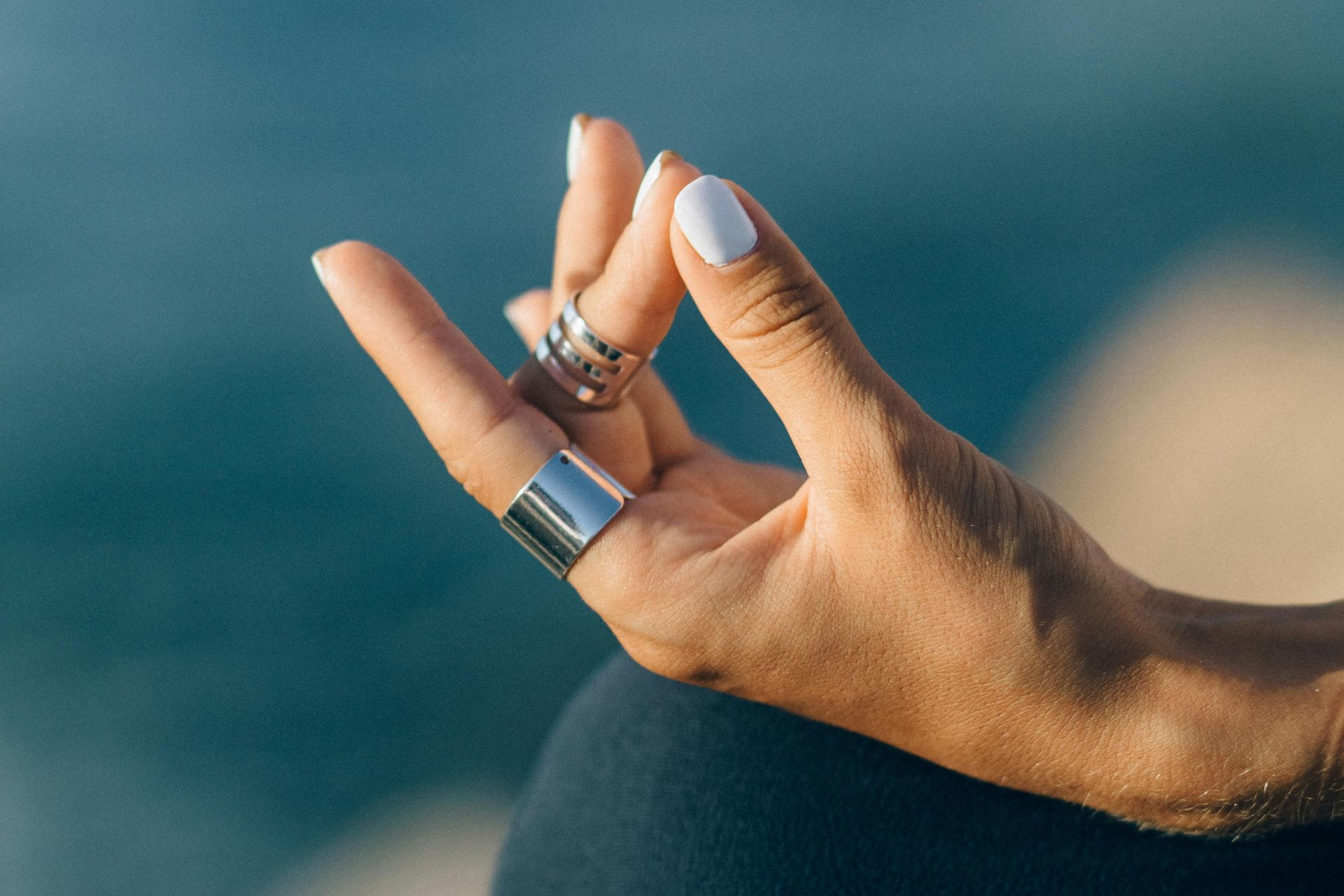 Hand with white nails, wearing silver rings, in a mudra position. Blue water blurred in the background.