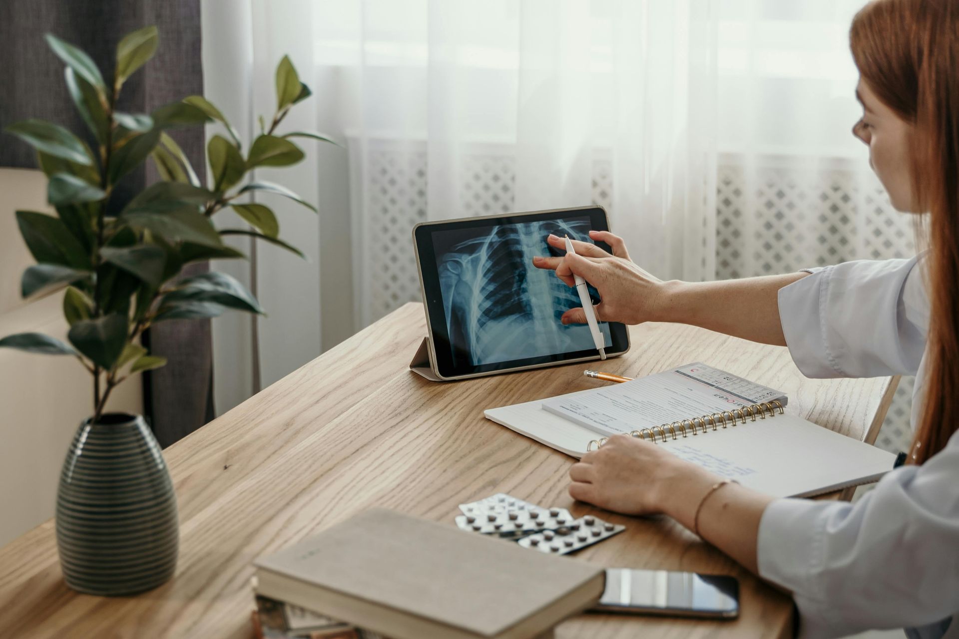 Doctor examining chest X-ray on tablet, pointing with stylus. Notebook, pills, and plant on desk.