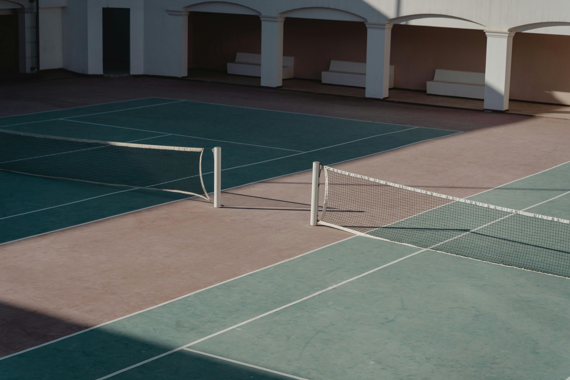 Tennis court with net, the surface divided into green and brown sections, under a covered area.