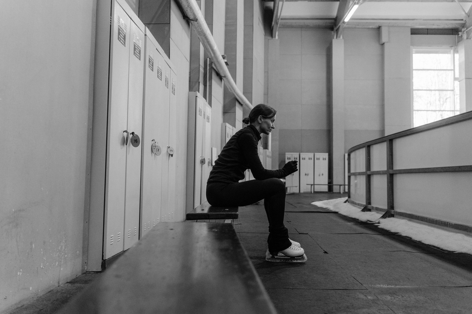 Woman in ice skates sits on a bench in an ice rink, looking at her phone.