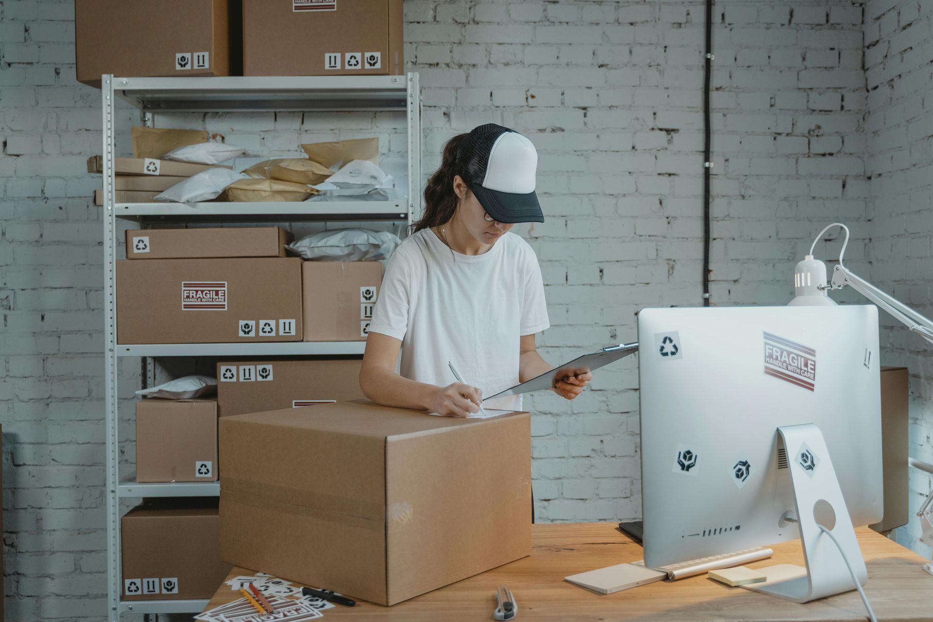 Woman in cap writing on clipboard at a desk with computer and boxes in warehouse.
