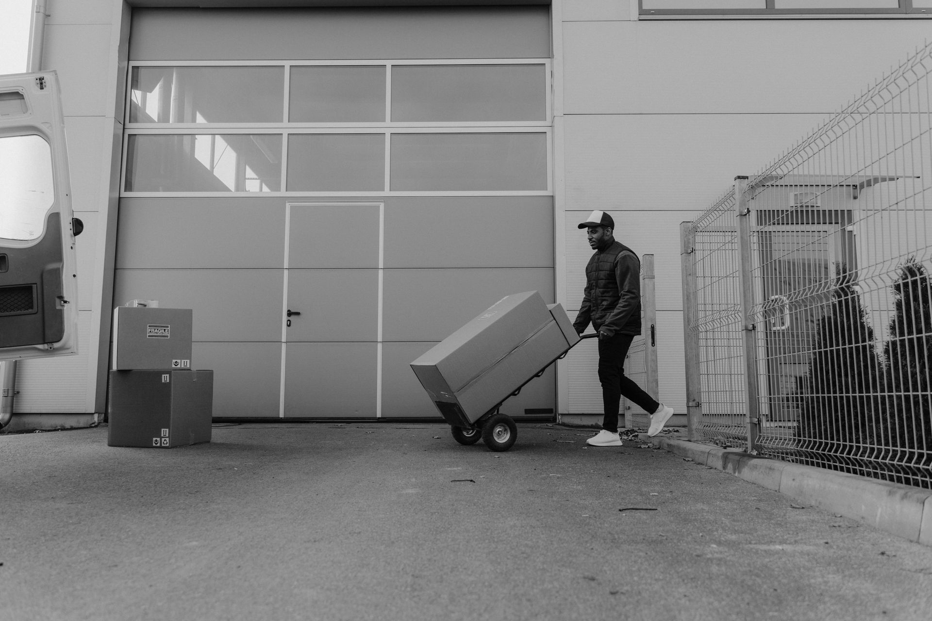 Man in jacket and hat pulls a large box on a hand truck near a loading dock.