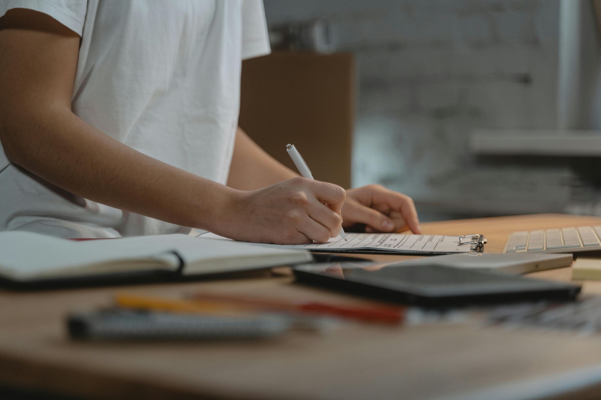 Person writing with a pen in a notebook on a desk with a keyboard and other office supplies.