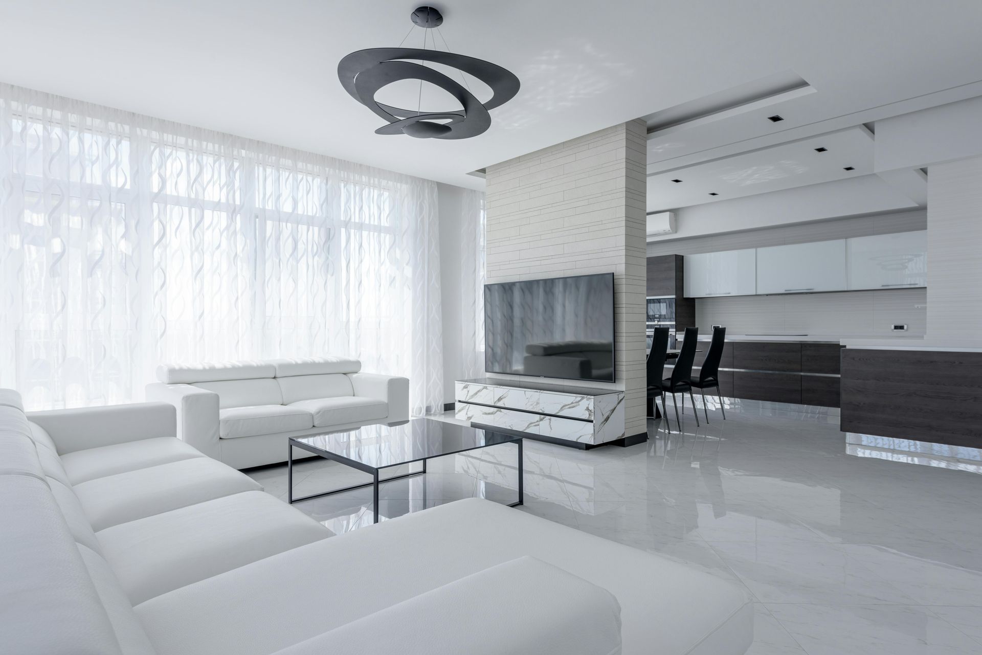 Bright, modern living room with white furniture, large windows, and an open kitchen area.
