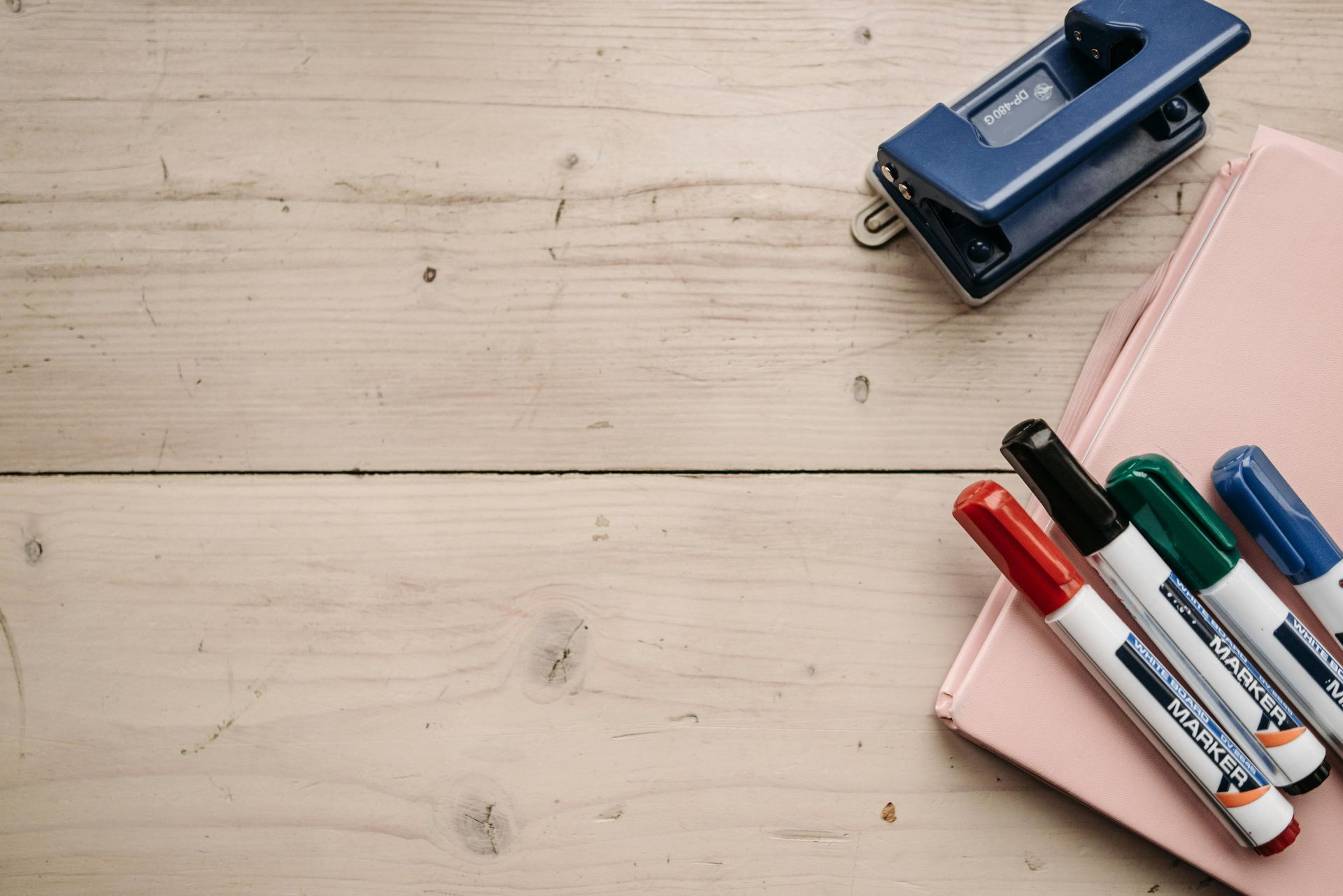 Wooden desk with a blue hole punch, pink notebook, and four colorful markers.