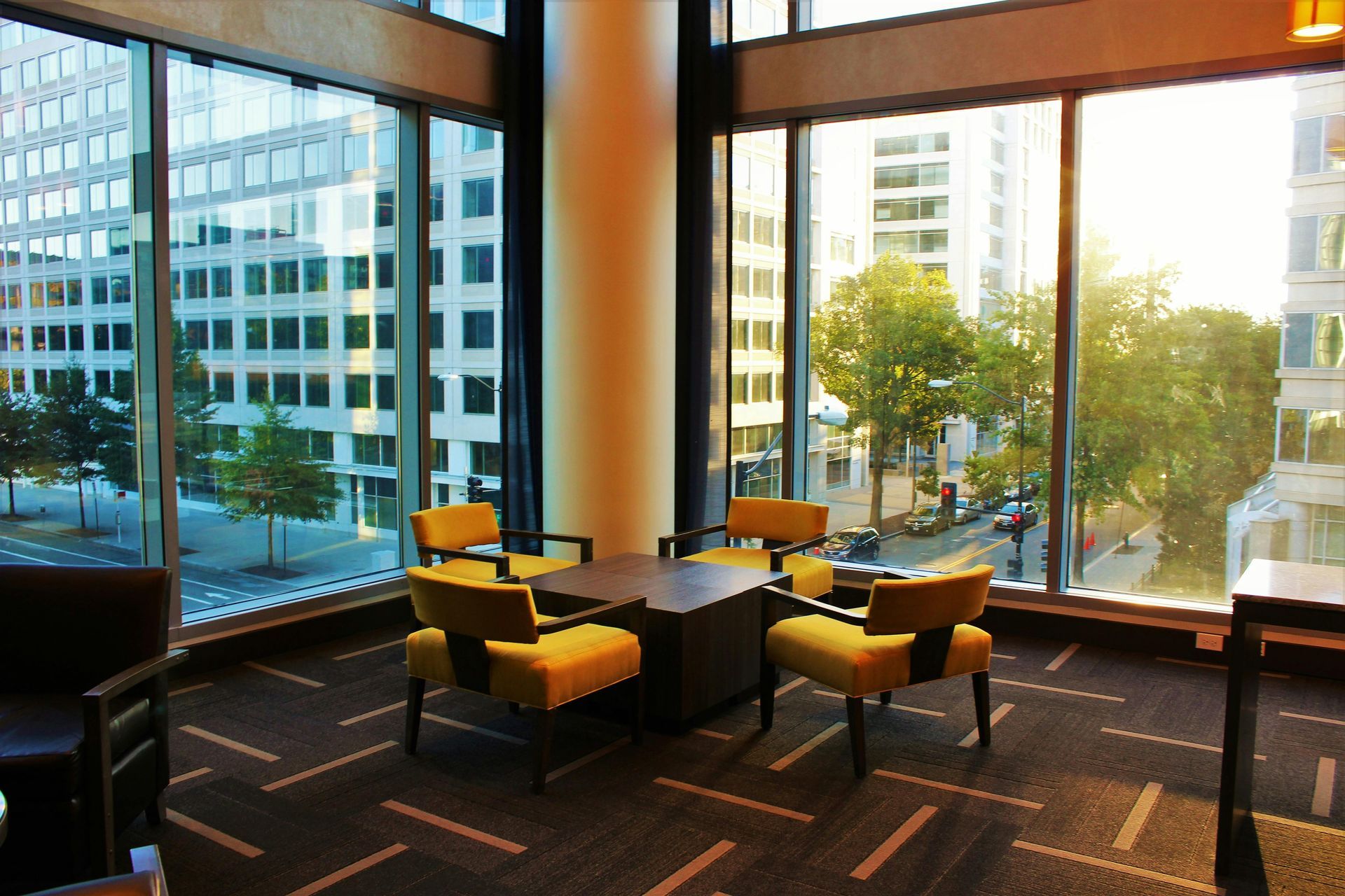 A modern office lounge with yellow chairs around a table, large windows, and city views.