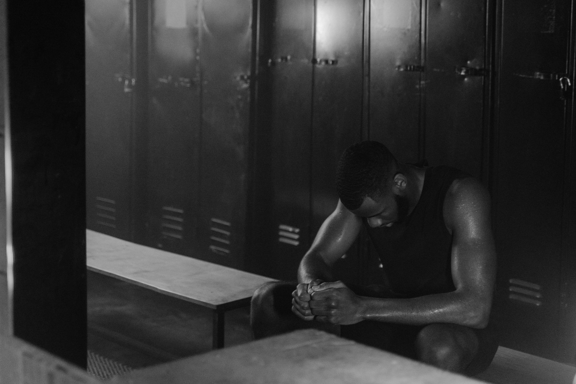 Man in a locker room, head down, hands clasped, sweaty, dark setting.