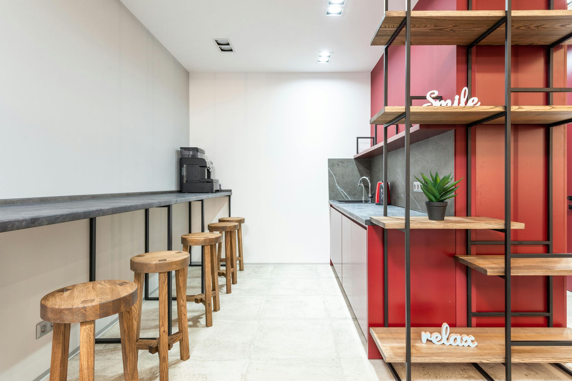 A modern office kitchen with red accents, wood shelves, a coffee machine, and bar stools.