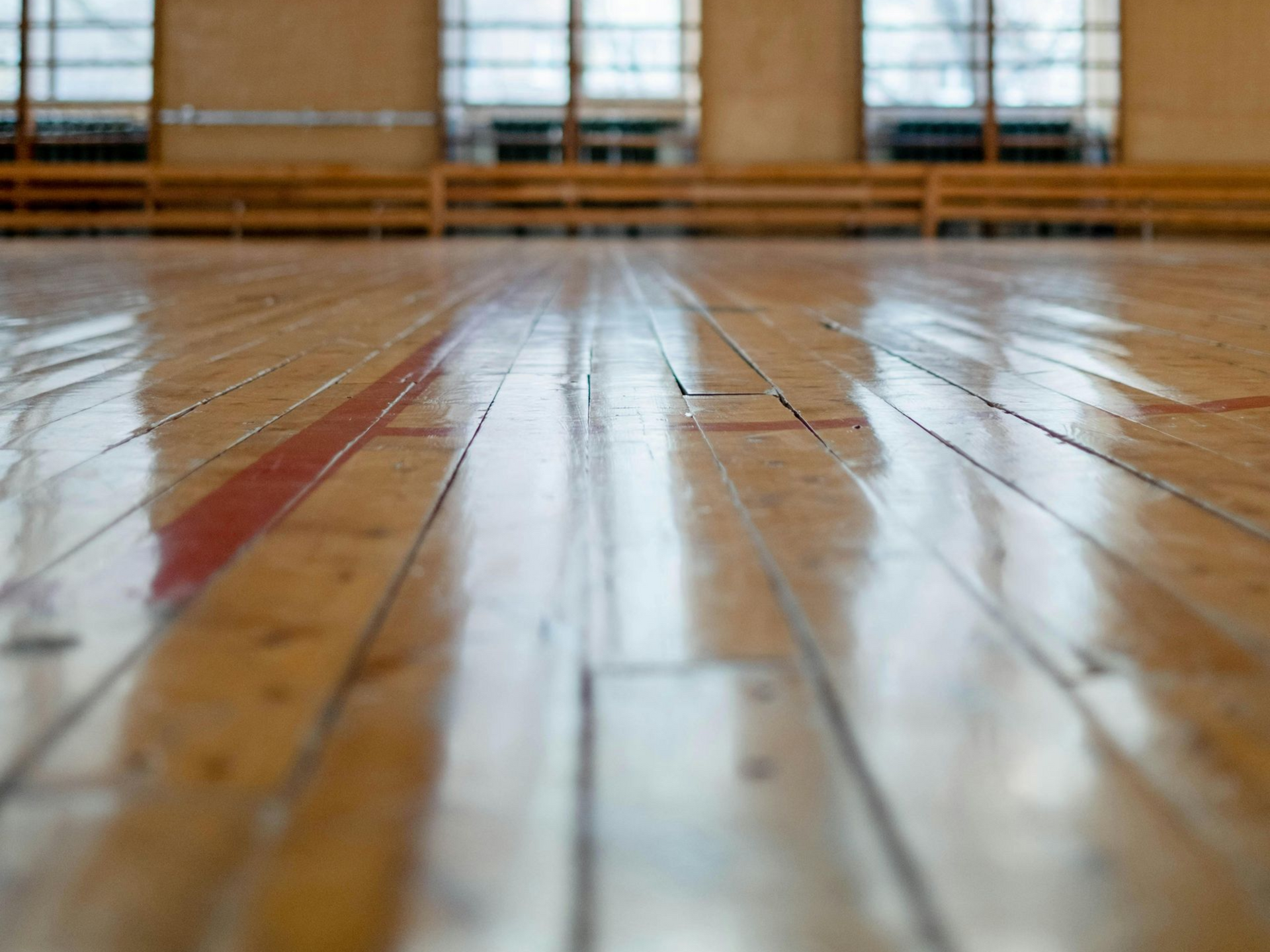 A low-angle view of a shiny, light-brown wood gym floor with red markings, facing windows in a bright indoor space.