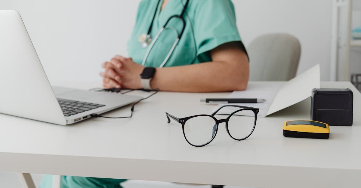 Doctor in green scrubs at a desk with laptop, glasses, and medical devices.