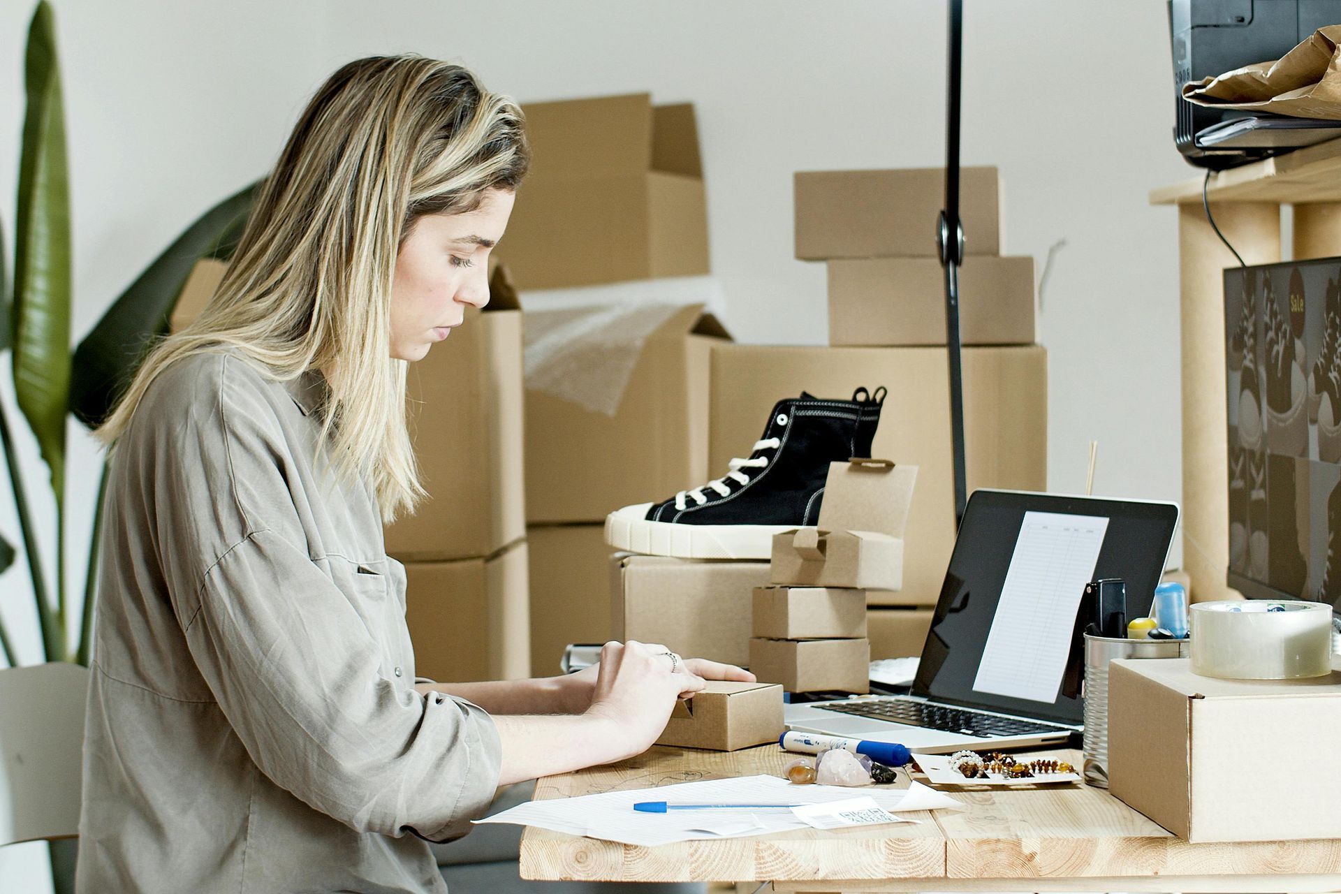 Woman packing boxes at a desk, surrounded by packages and a laptop.