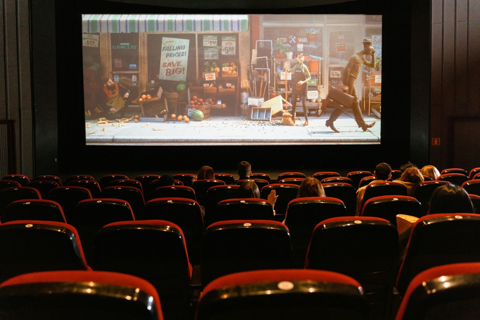 Audience watches a movie in a dark theater. Red seats face a large screen with a film scene.