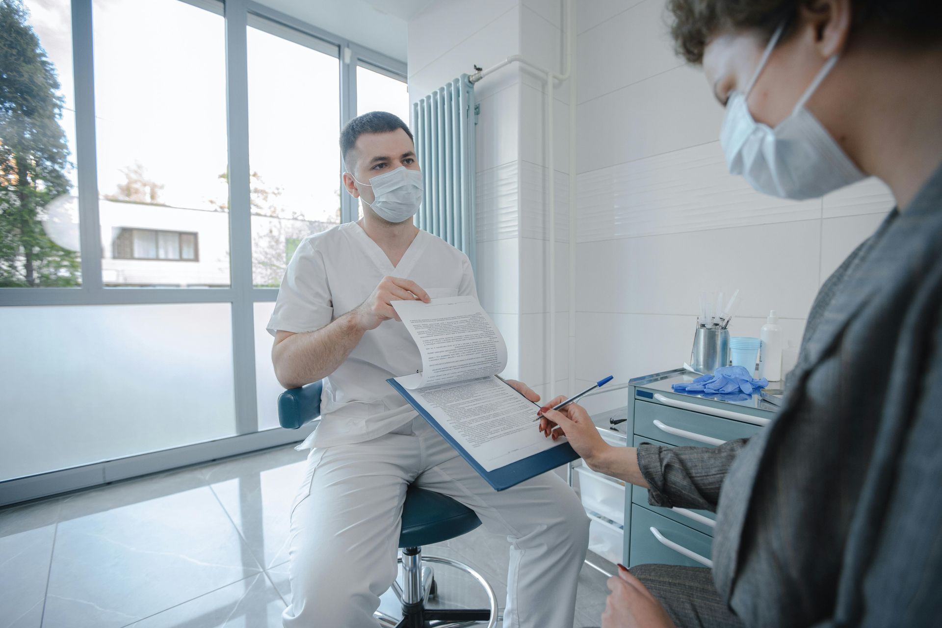 A medical professional in a mask and white scrubs shows a patient paperwork in an exam room.