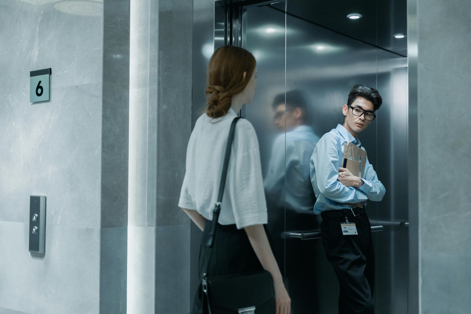 Woman and man in an elevator. The man looks at the woman, holding a tablet. Gray interior.