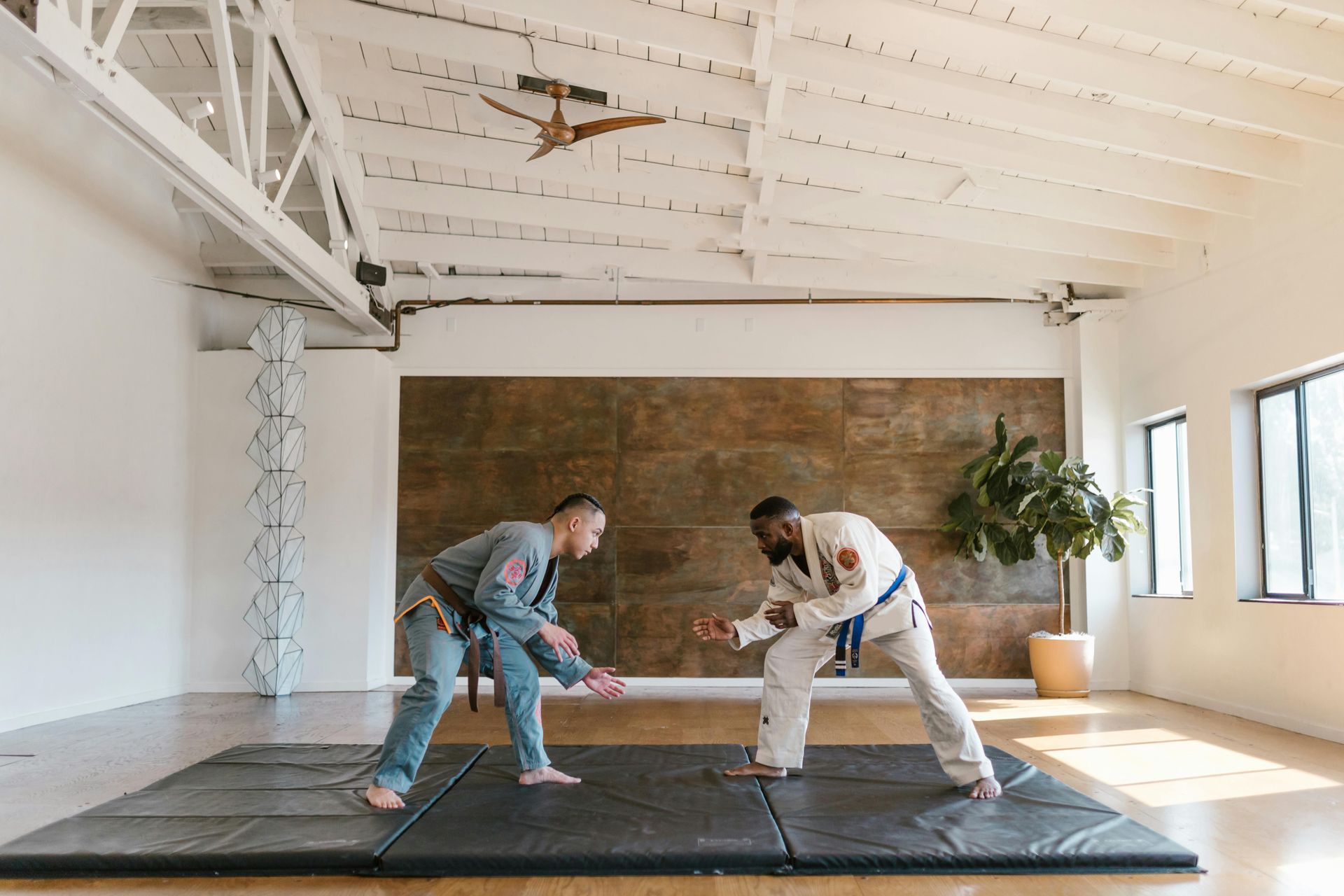 Two men in martial arts uniforms face each other on a mat, preparing to spar indoors.