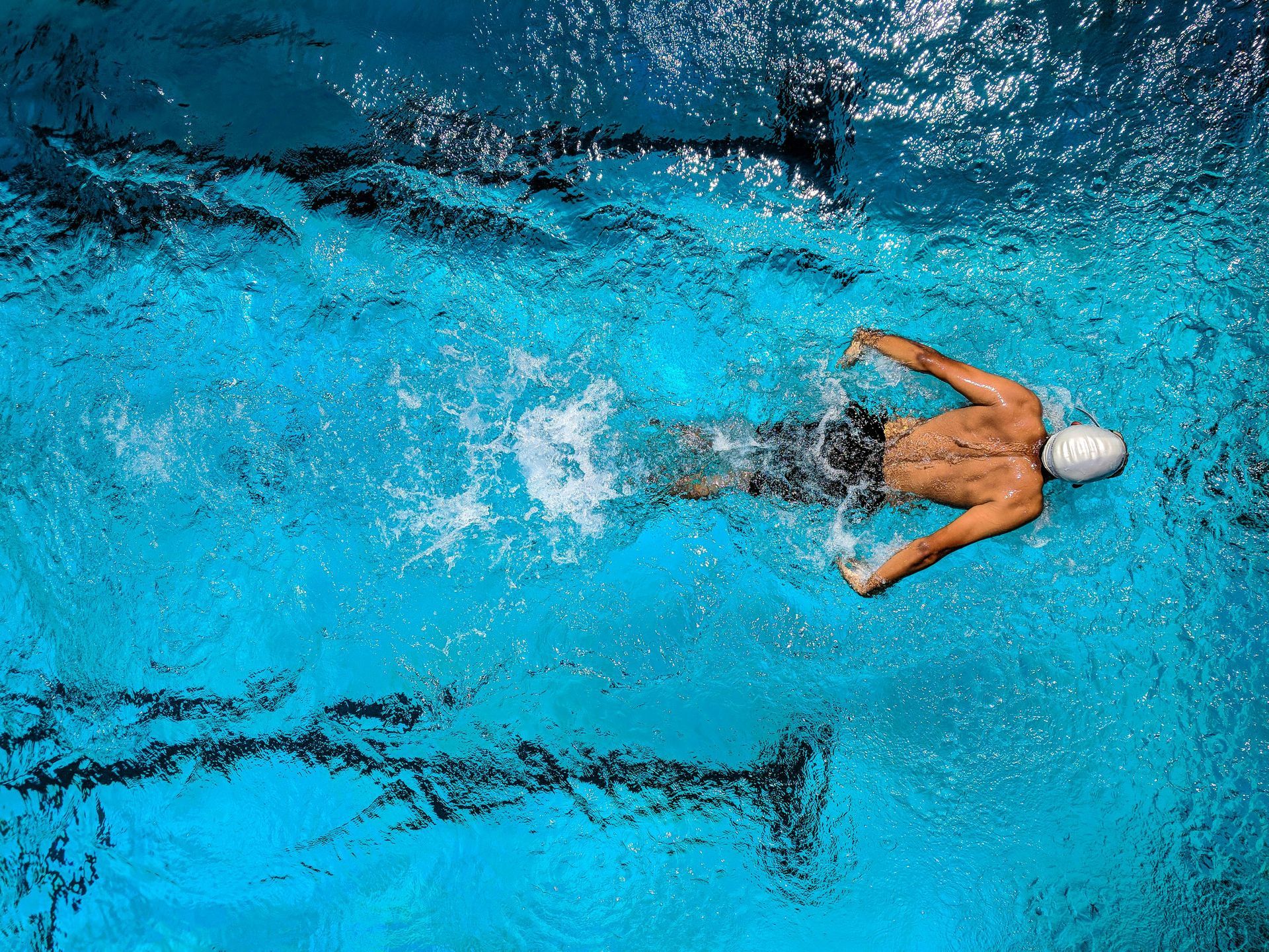 Swimmer in white cap, stroking through a blue pool, viewed from above.