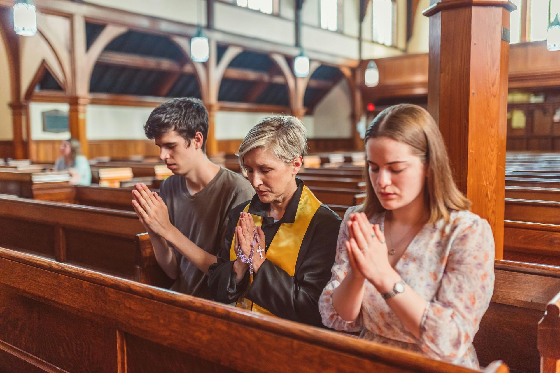 Three people praying in a church, hands clasped. Two young adults, one older woman, all with closed eyes.