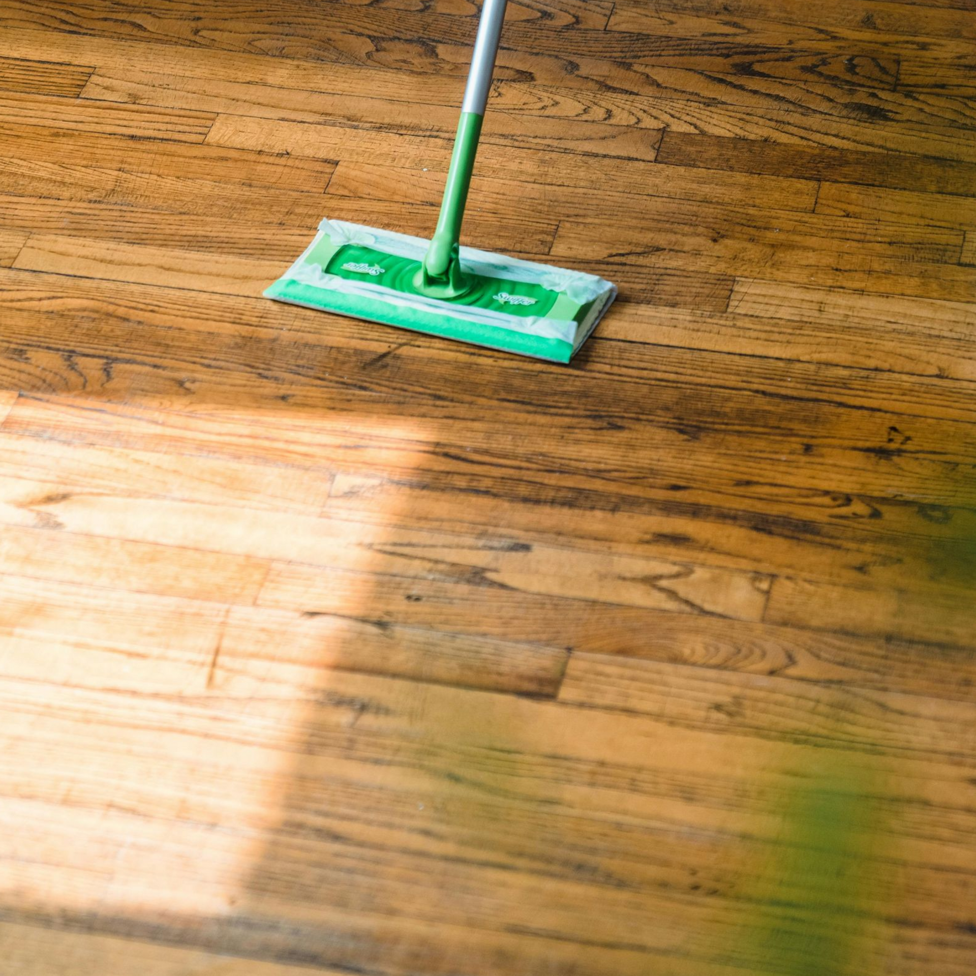 A green flat mop cleaning a light brown hardwood floor.