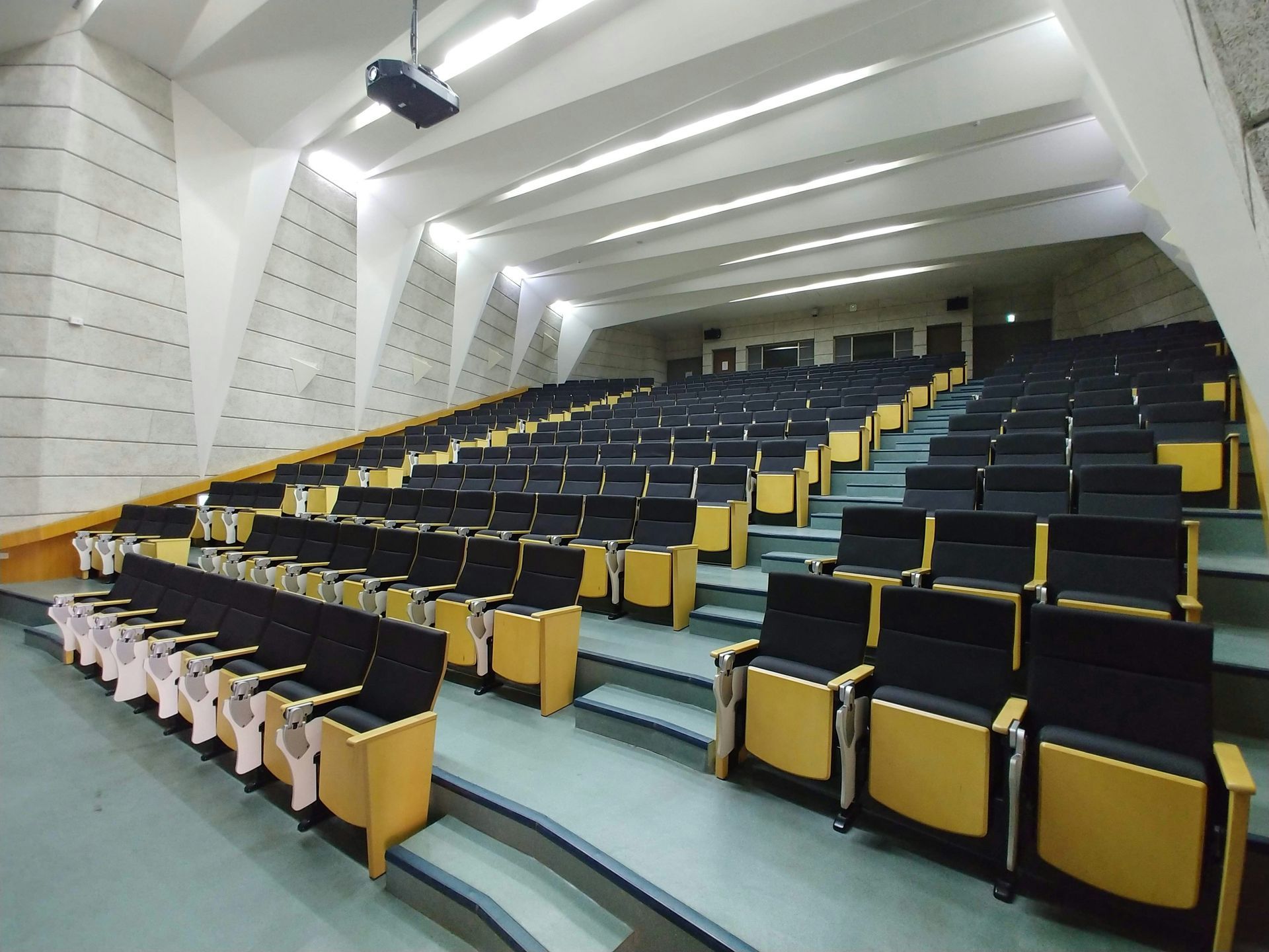 Auditorium with rows of black seats on stepped levels; beige seat backs; sloped, patterned walls.
