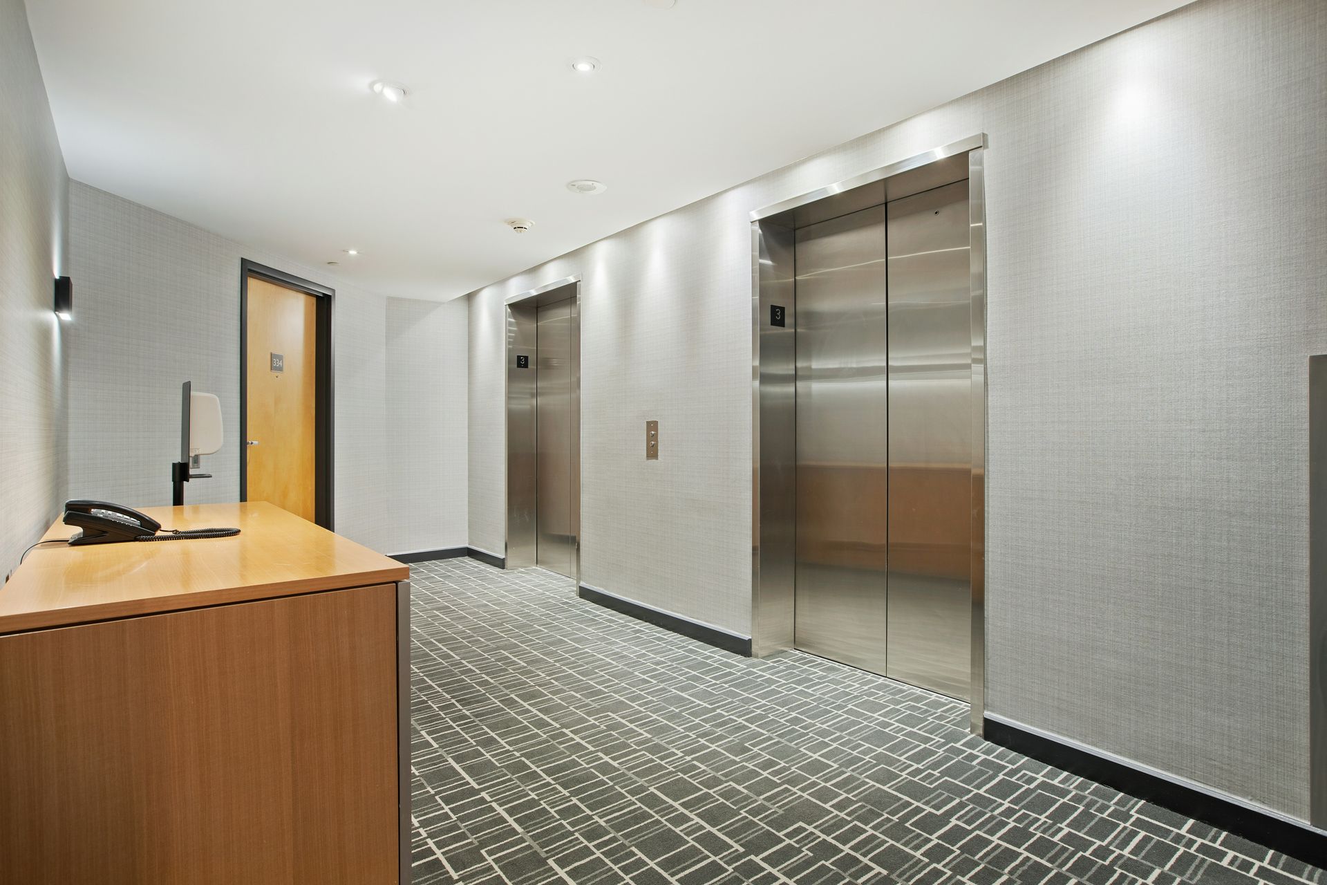 Hallway with two elevators, a wooden counter, and a door. Gray walls and carpet.