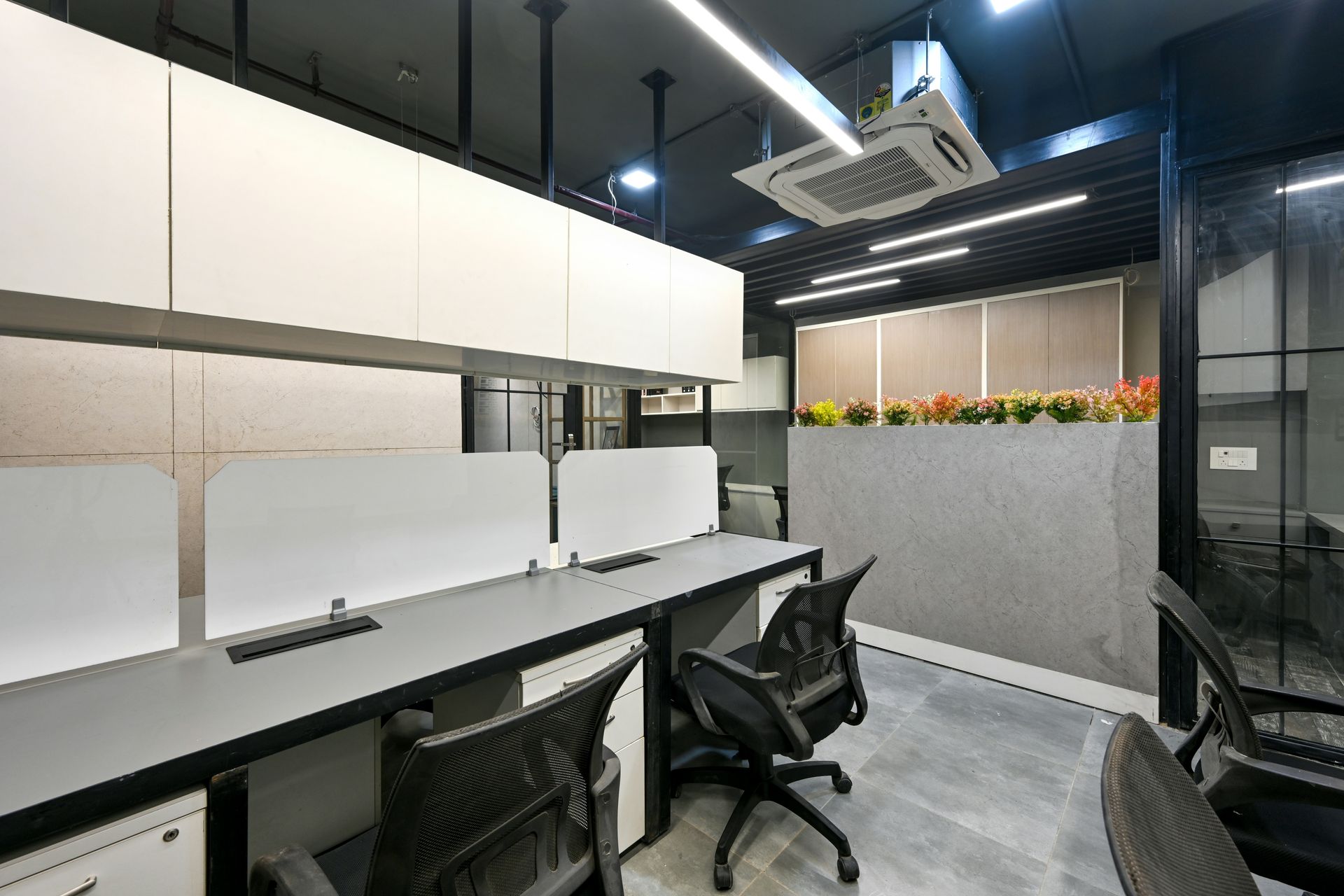 Office workspace with desks, chairs, and dividers, with overhead storage and a plant wall.