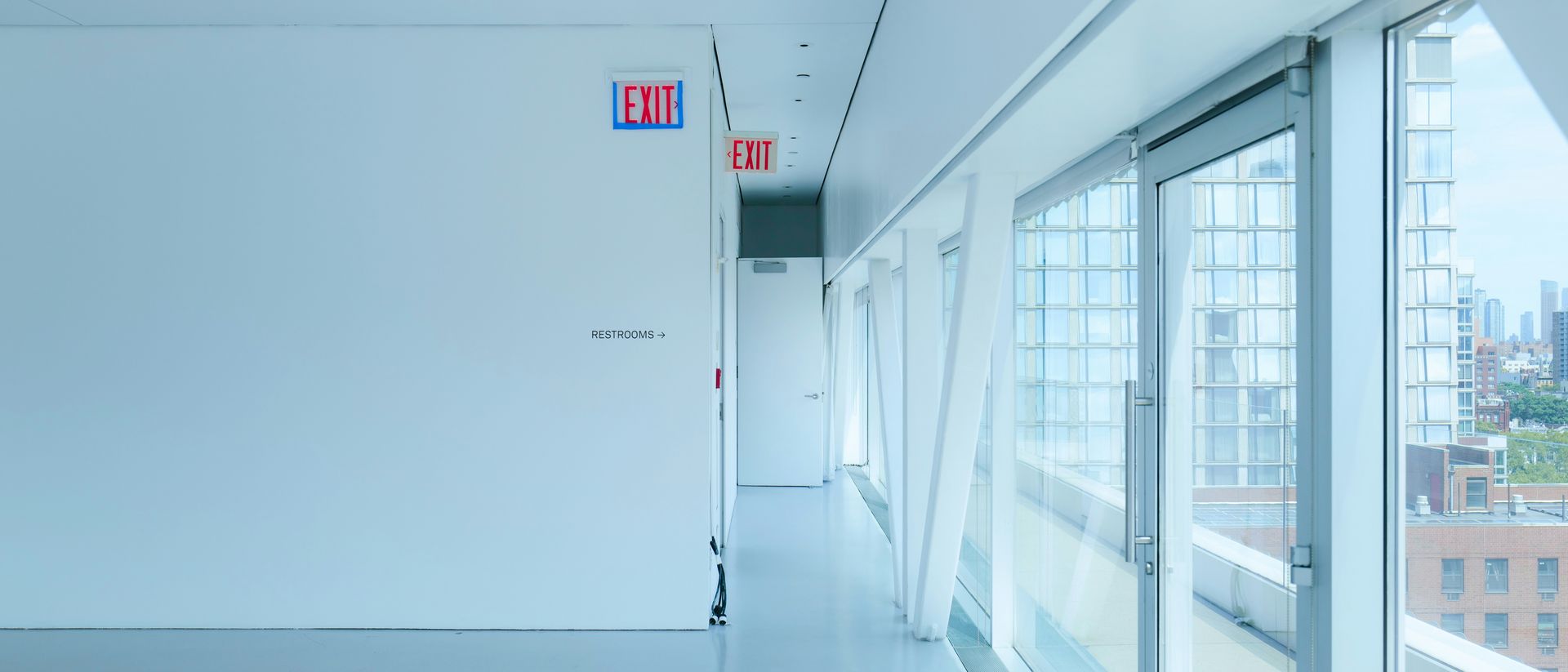 Bright, white hallway with large windows overlooking a city, an exit sign above a door.