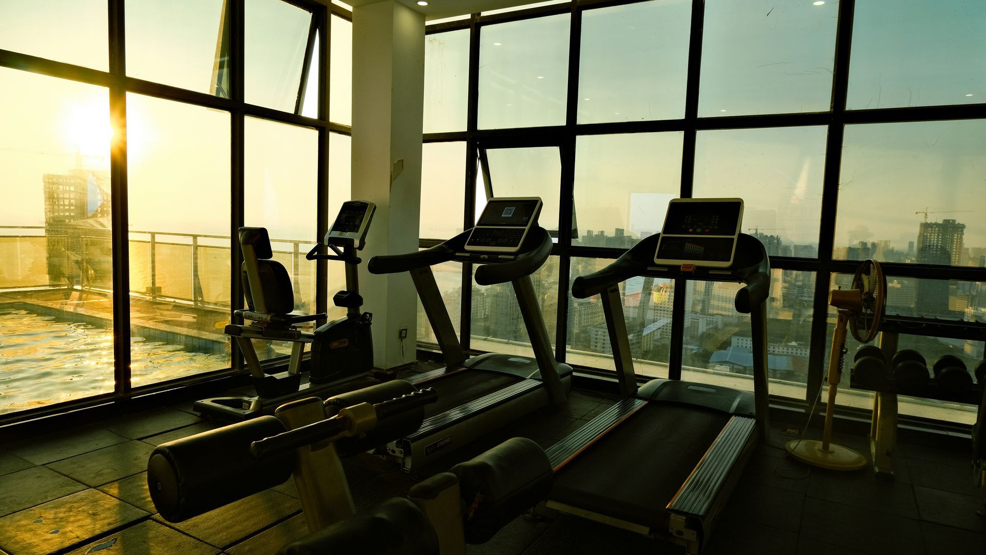Gym with treadmills, stationary bike, and weights, offering city views through large windows at sunset.