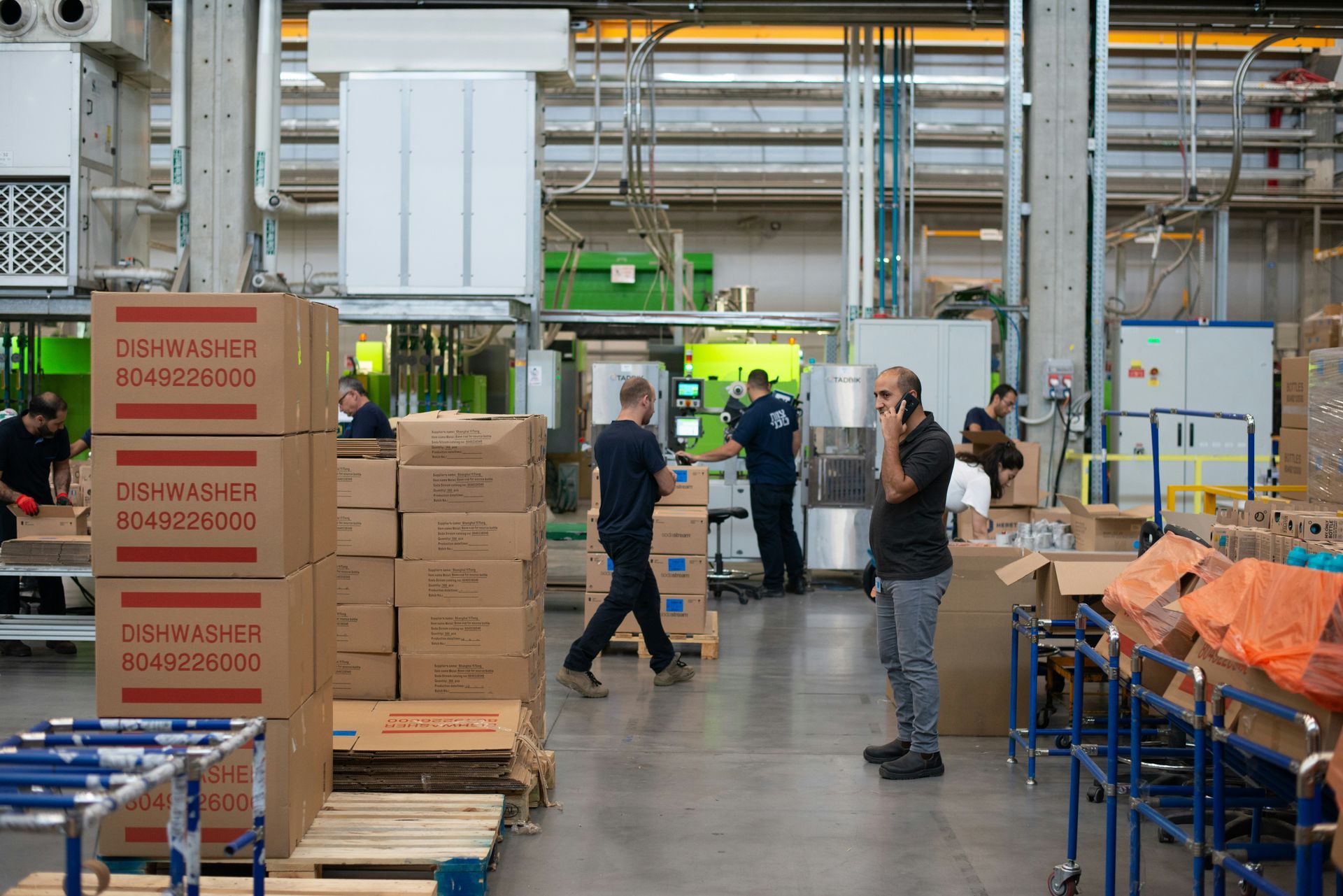 Warehouse interior with people working. Boxes, machinery, and workers are visible.