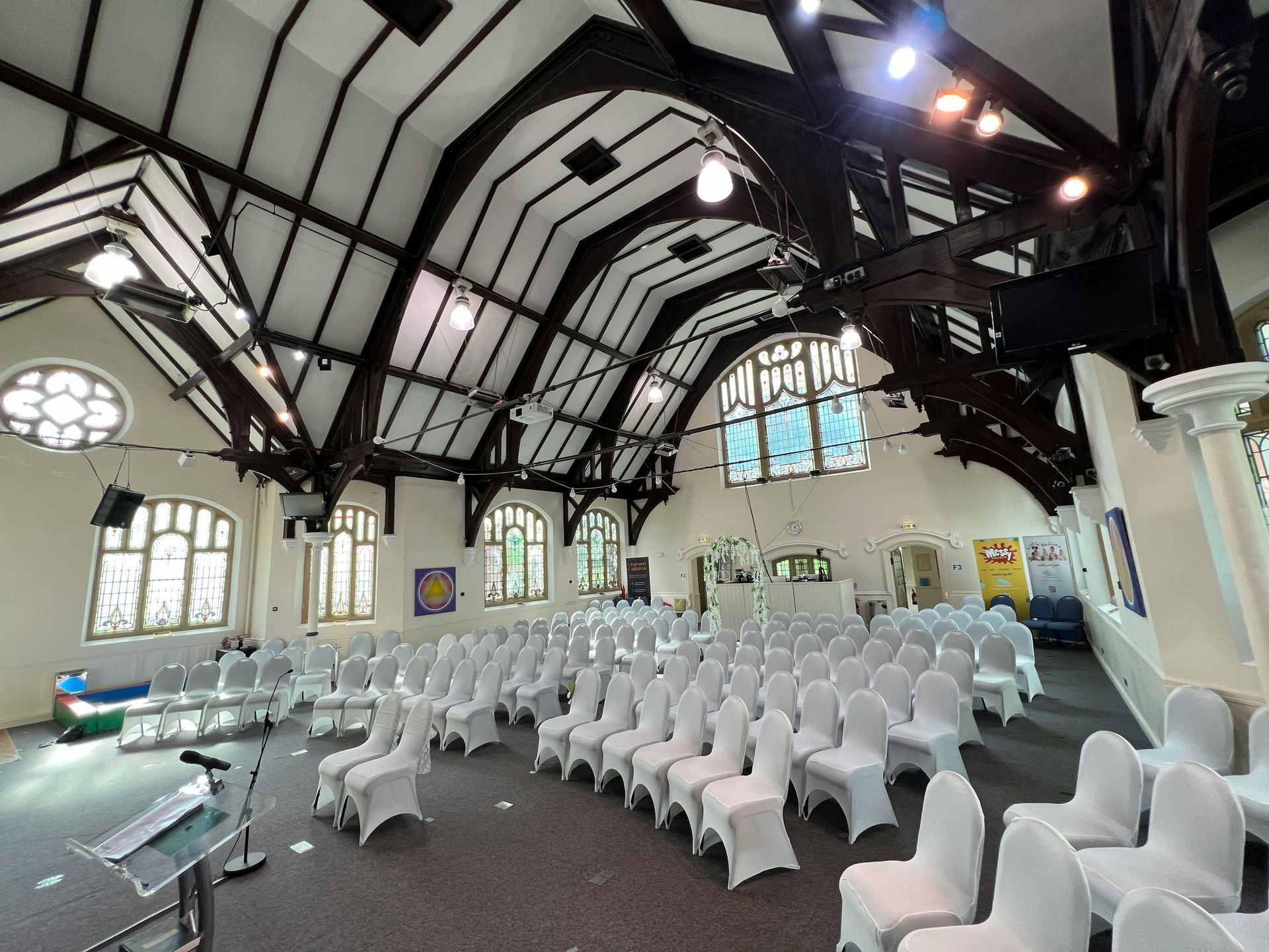 Interior of a chapel prepared for an event, with rows of white-covered chairs, a podium, and arched windows.