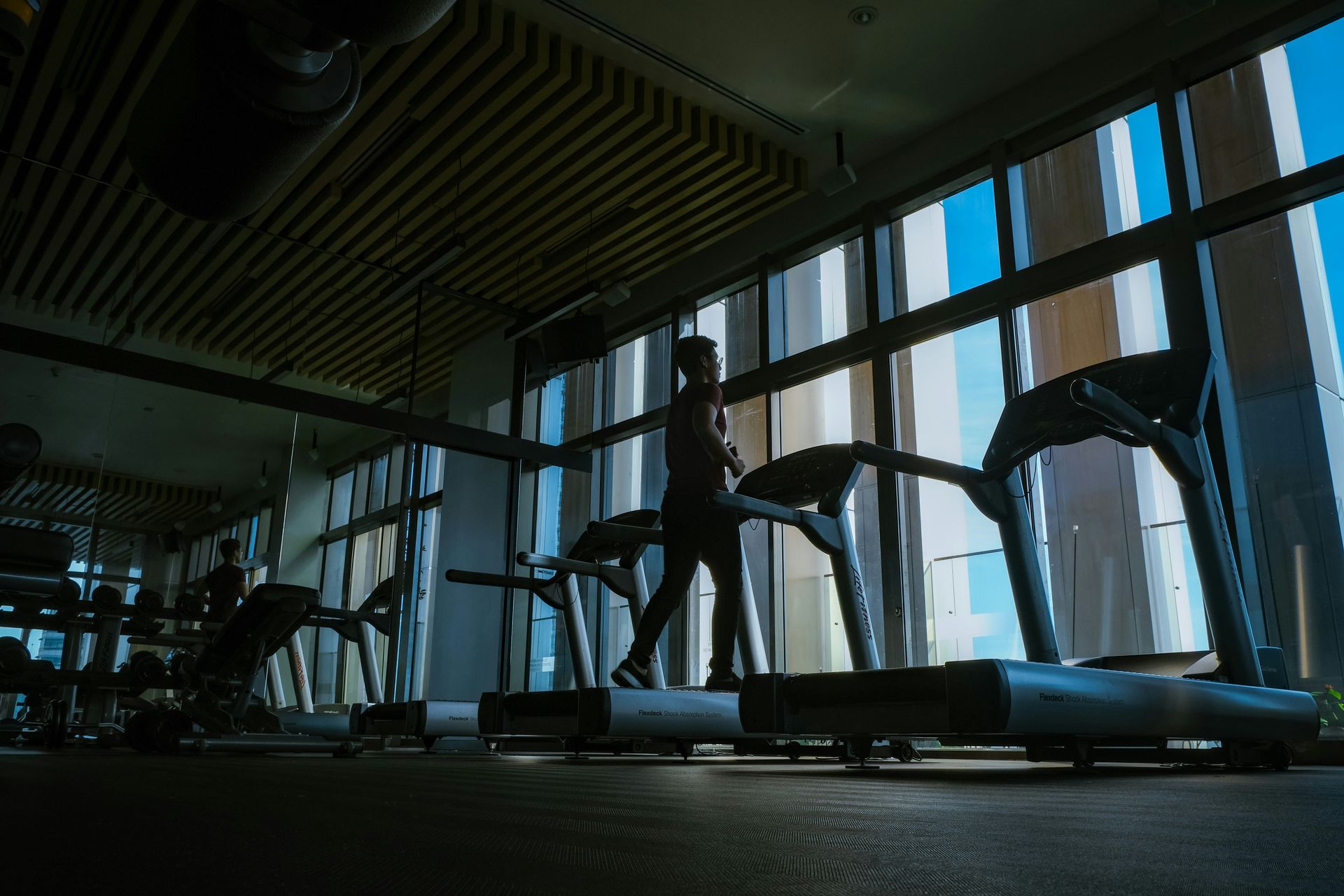 Person walking on a treadmill in a gym with large windows; silhouette against bright blue sky.