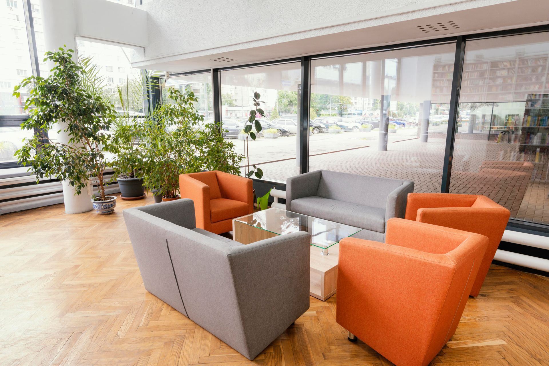 A modern lounge area with gray and orange seating around a glass table, next to large windows and plants.