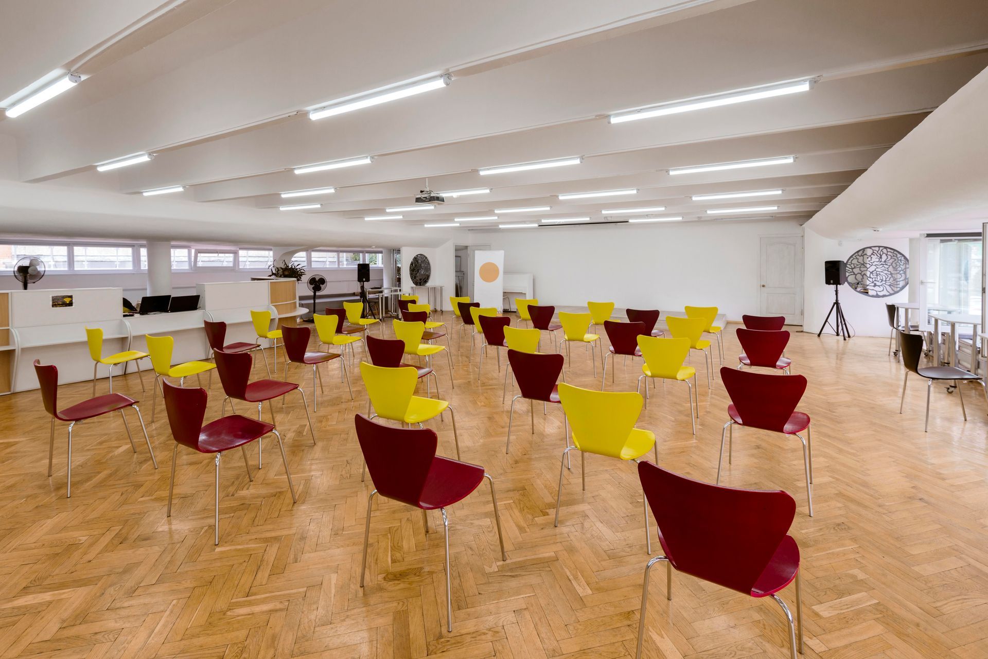 Auditorium with rows of red and yellow chairs; a blank white wall and wooden floor.