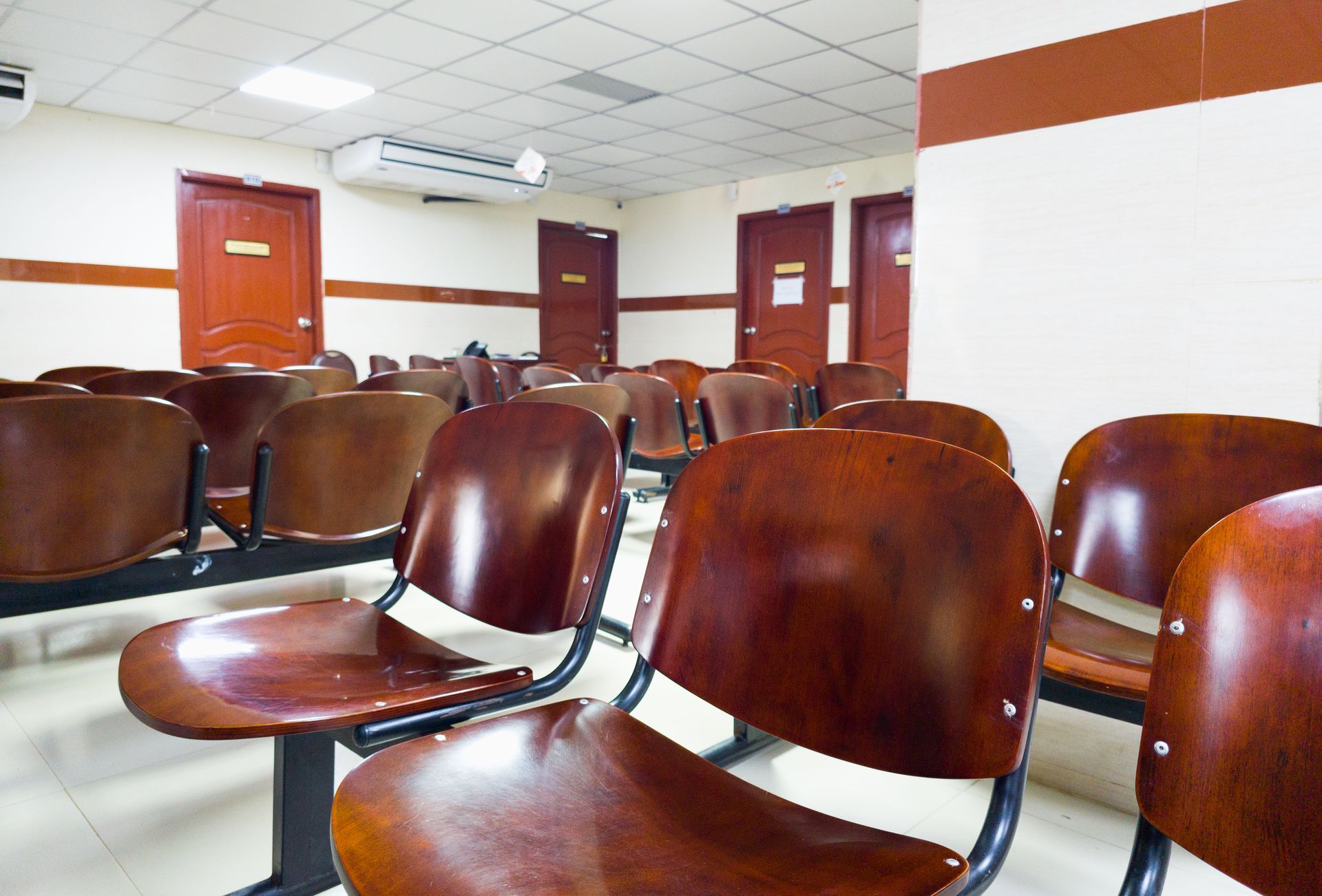 Rows of brown wooden chairs in a waiting room, with doors and air conditioning visible.