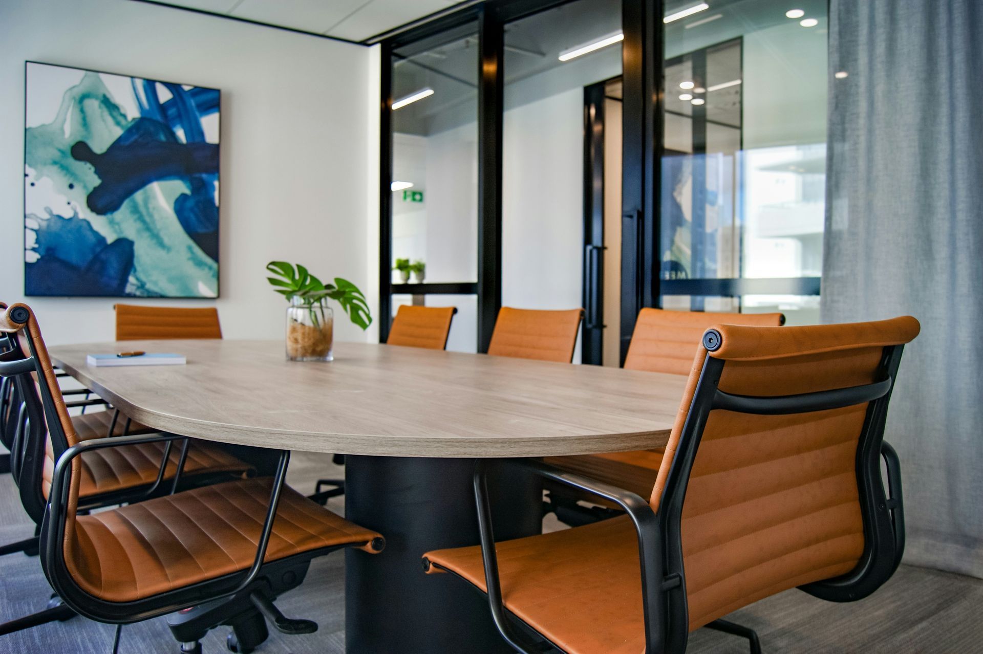 Conference room with a light wood oval table, tan leather chairs, and blue abstract art.