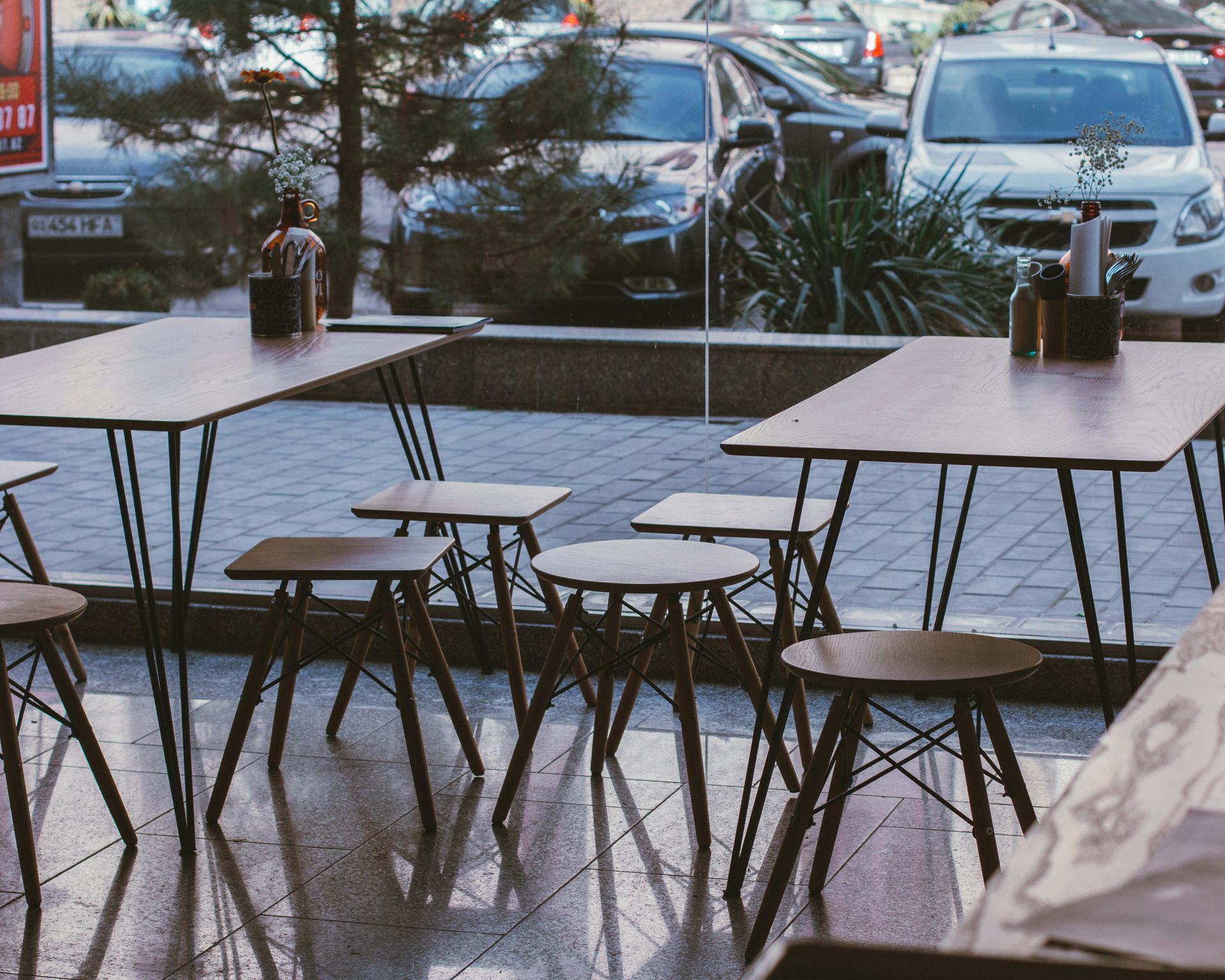 Restaurant tables and stools inside, with reflections on the floor, overlooking a street with parked cars.