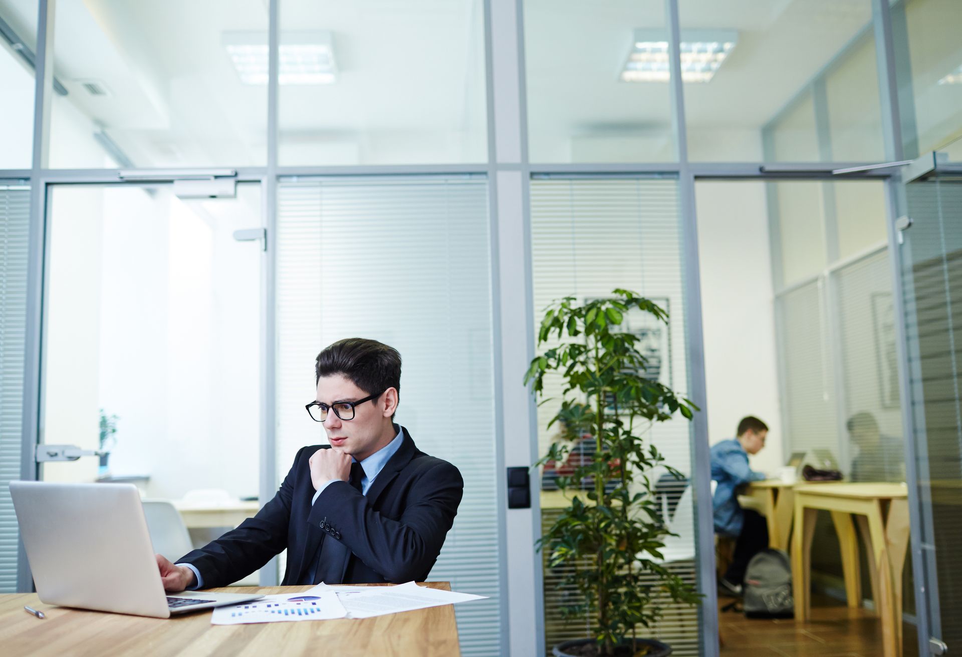 Man in suit with glasses looking at a laptop in a modern office, other people in the background.