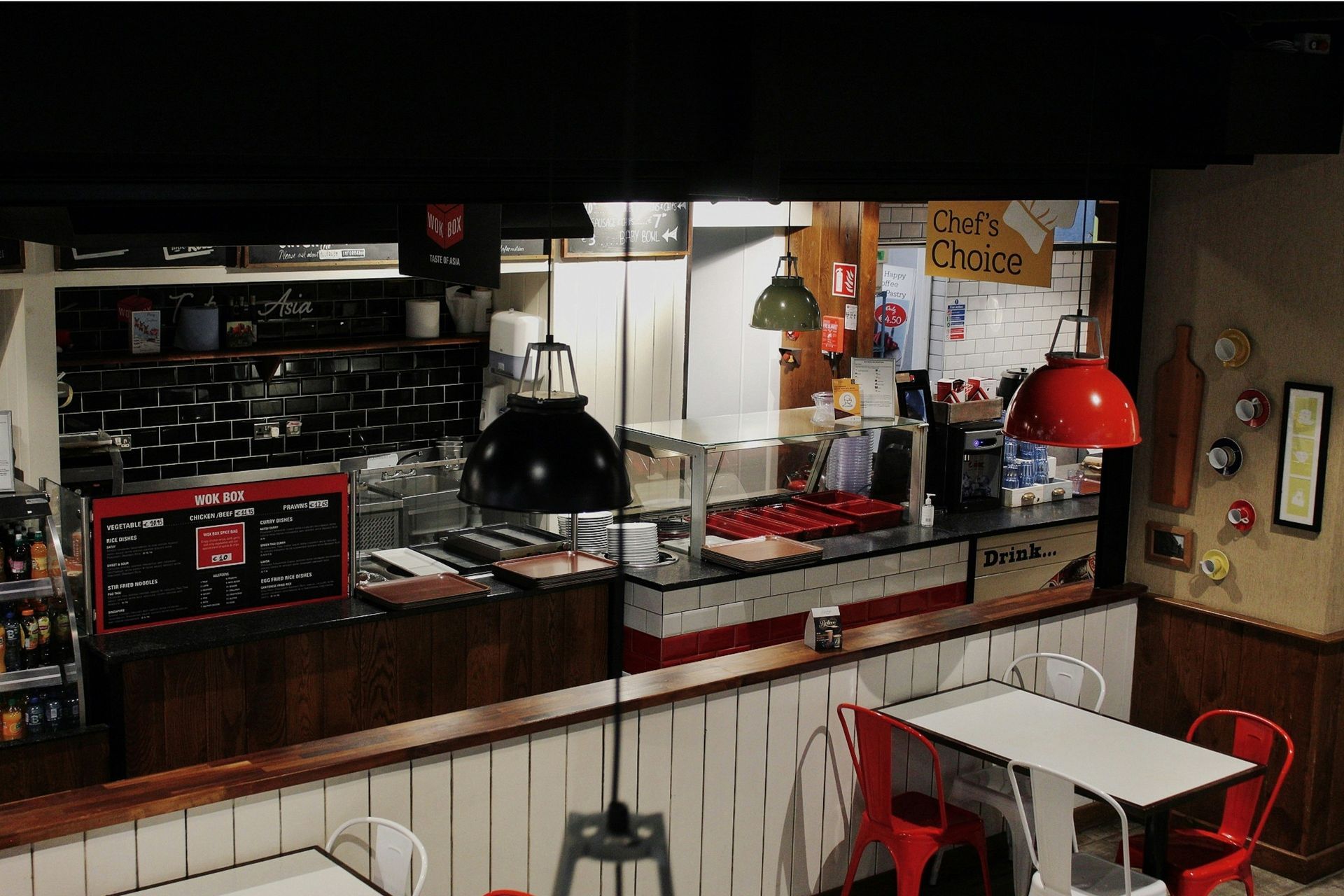 Restaurant interior: counter with food prep area, red chairs, white tables. Dark wood, tiled wall, and hanging lights.