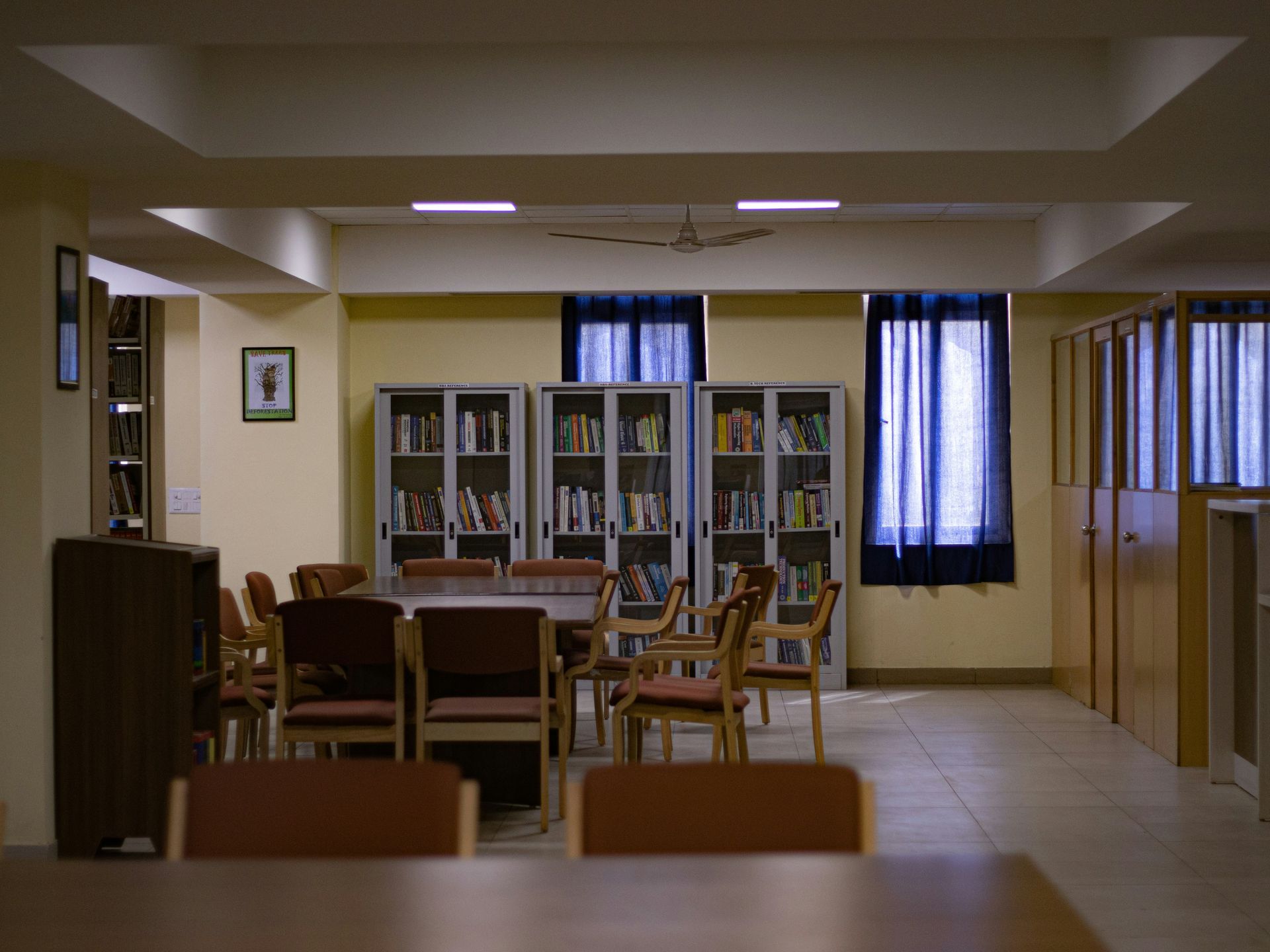 Library interior with tables, chairs, bookshelves, and windows with dark blue curtains.