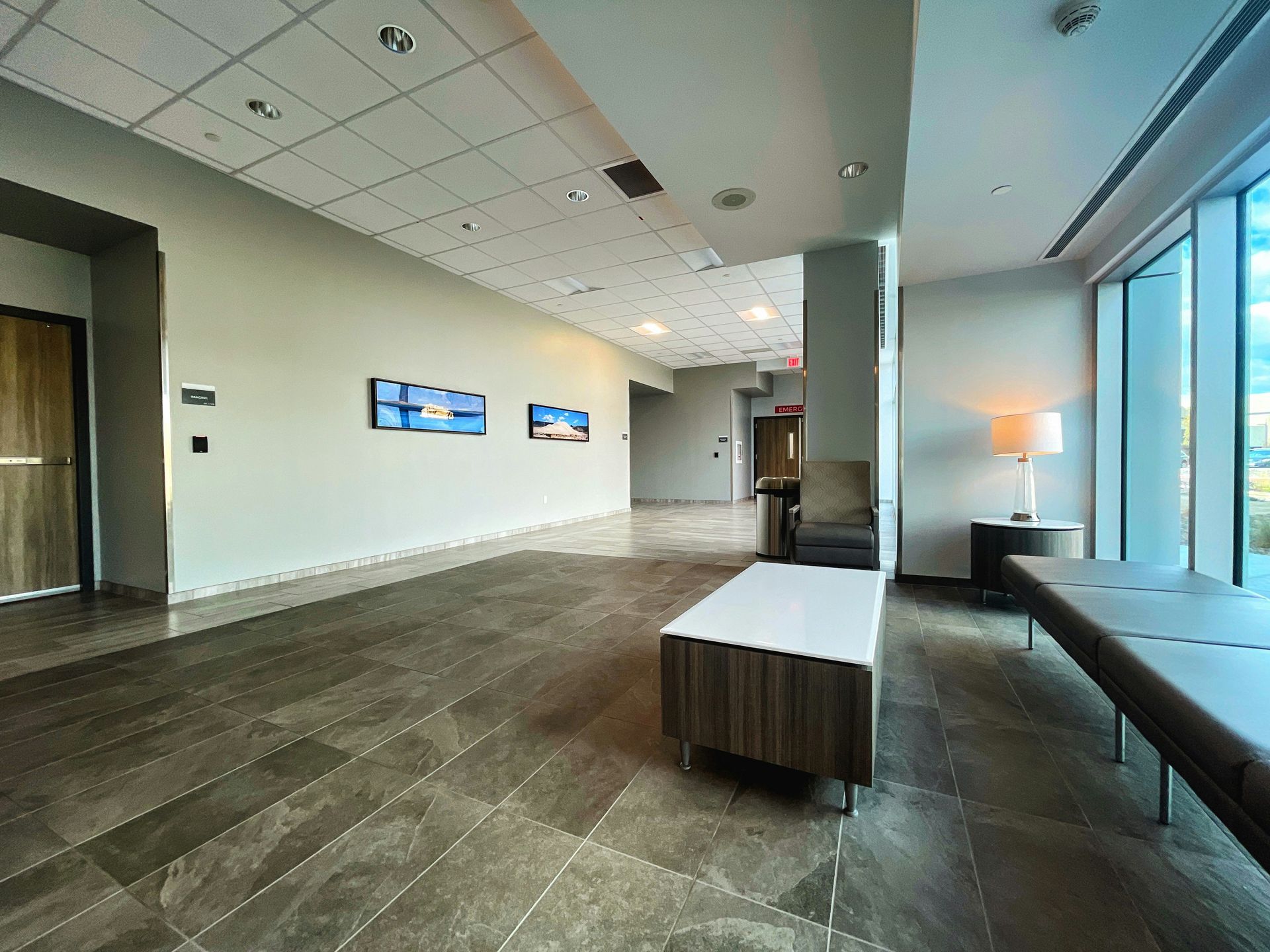 Lobby with a long bench, artwork, and large windows. Gray tile floor, beige walls, and natural light.