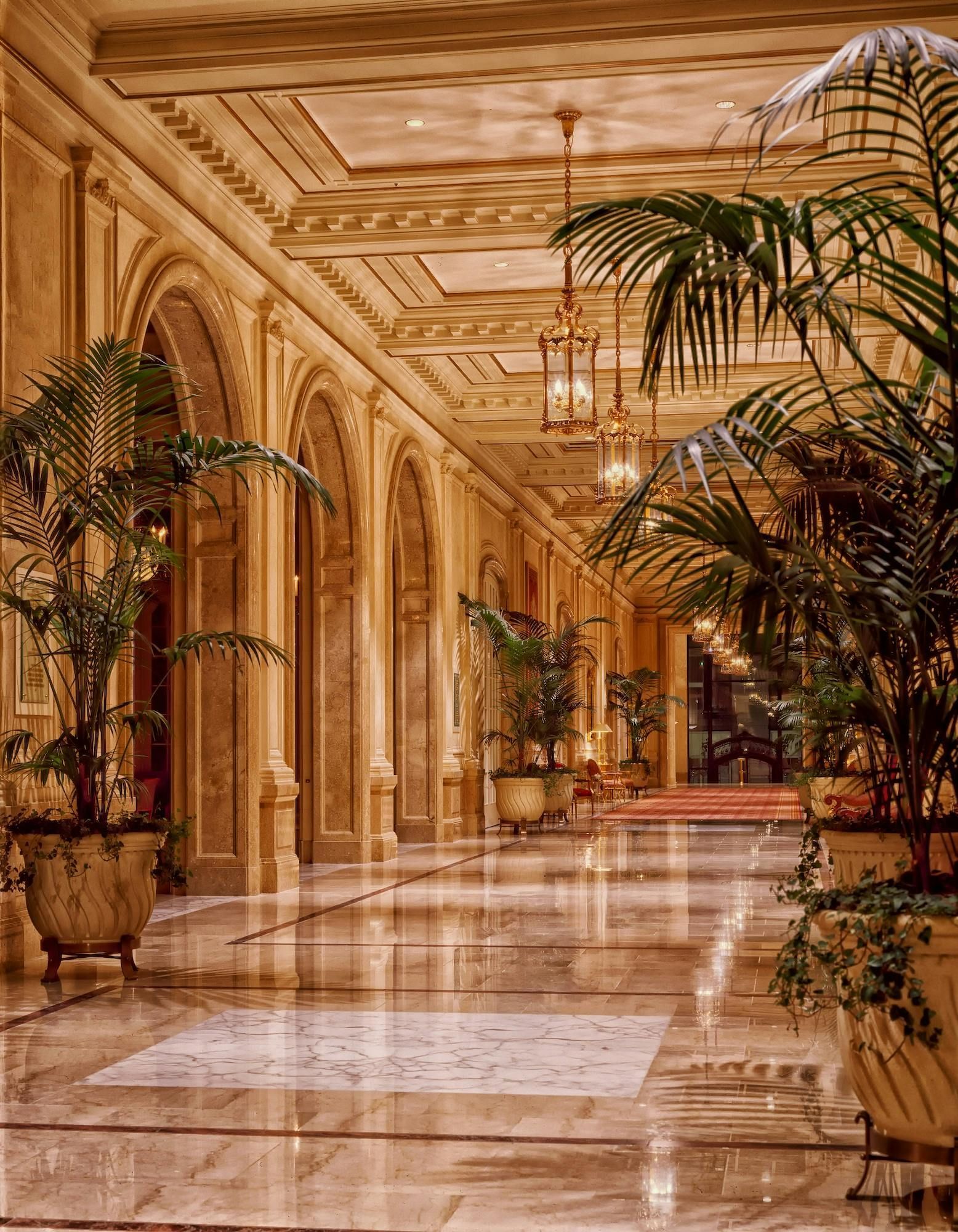 Elegant hallway with arched doorways, marble floors, potted plants, and chandeliers.